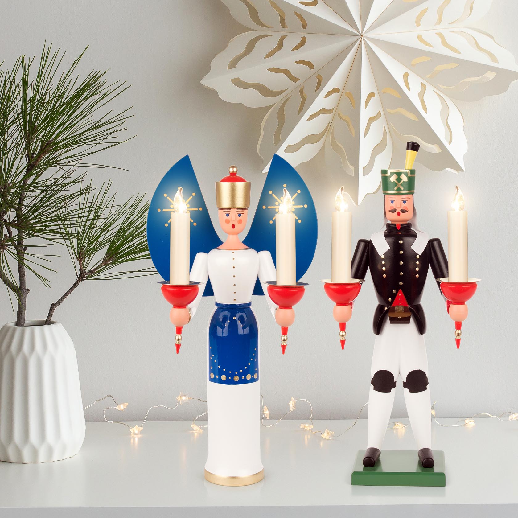A wooden angel figure with blue wings holding two candles stands next to a soldier figurine with red candle holders.