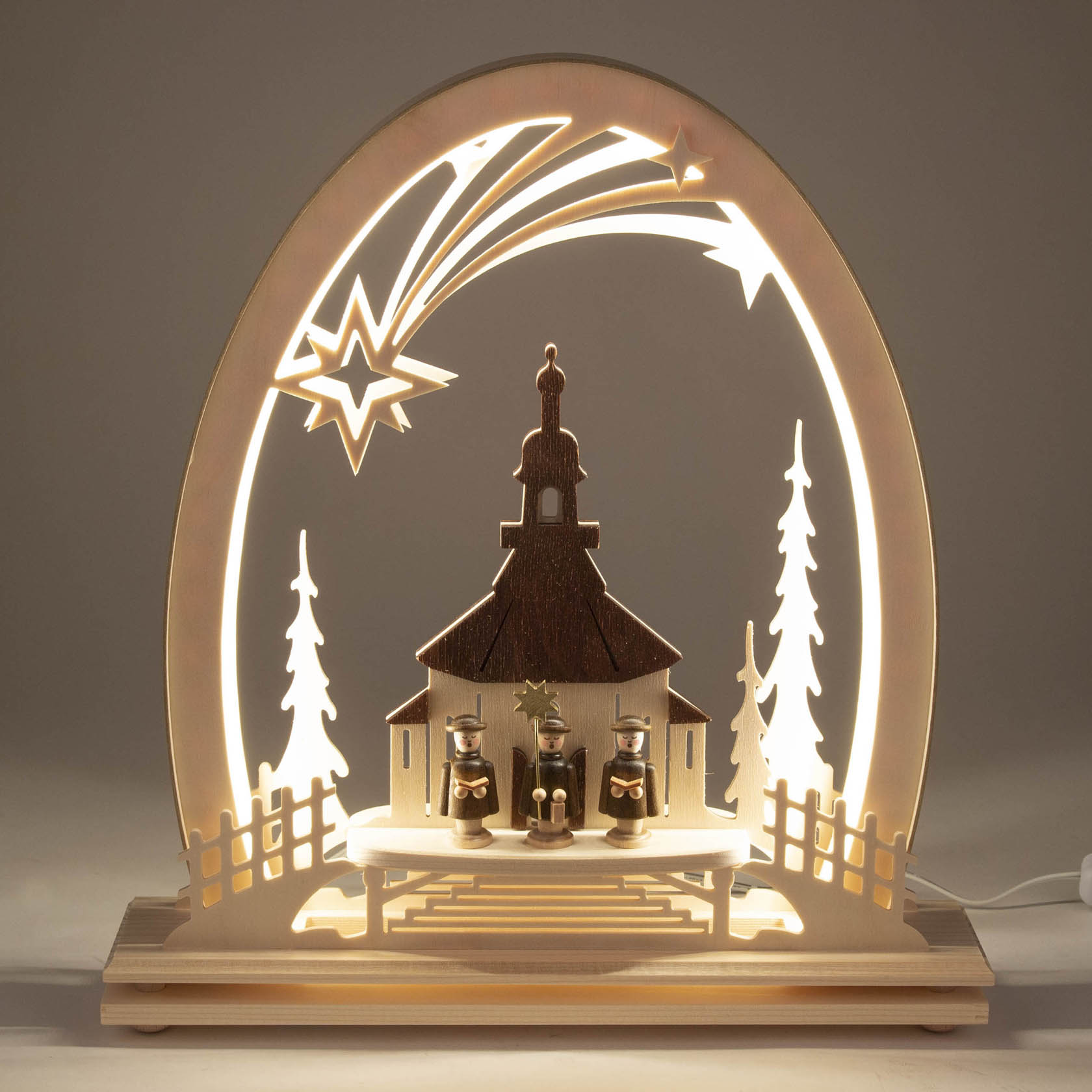 Erzgebirgsbogen mit beleuchteter Seiffener Kirche, flankiert von zwei Bäumen. Drei Kurrende-Sänger stehen vor der Kirche unter einem hellen, leuchtenden Sternbogen.
