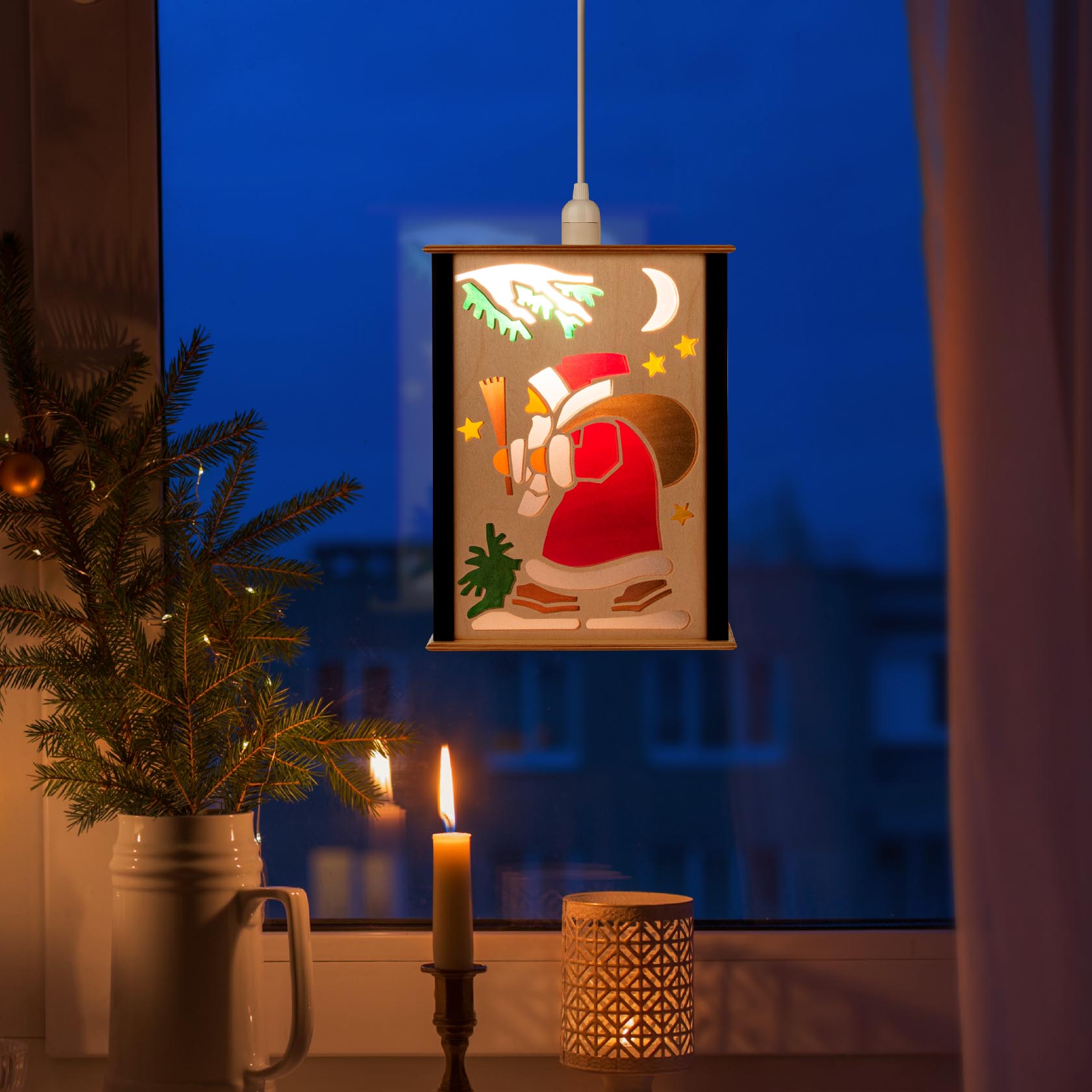 A lit paper lantern hanging in a window, featuring a child-like Santa Claus carrying a sack and lantern with stars and a crescent moon.