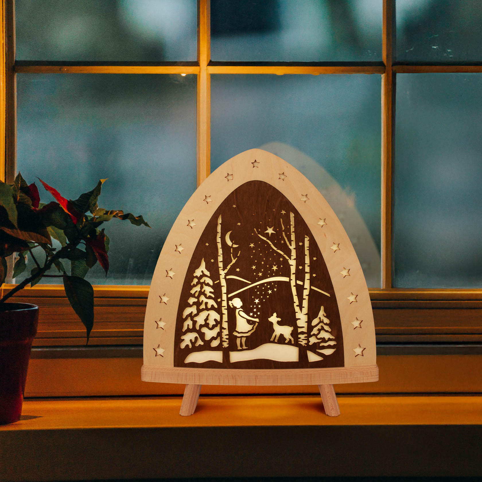 Holz-Dekorationsleuchte mit ausgestanztem Winterwaldmotiv, zeigt ein Rentier im Schnee unter einem Sternenhimmel; von kleinen Sternen umrahmt, auf Fensterbank.
