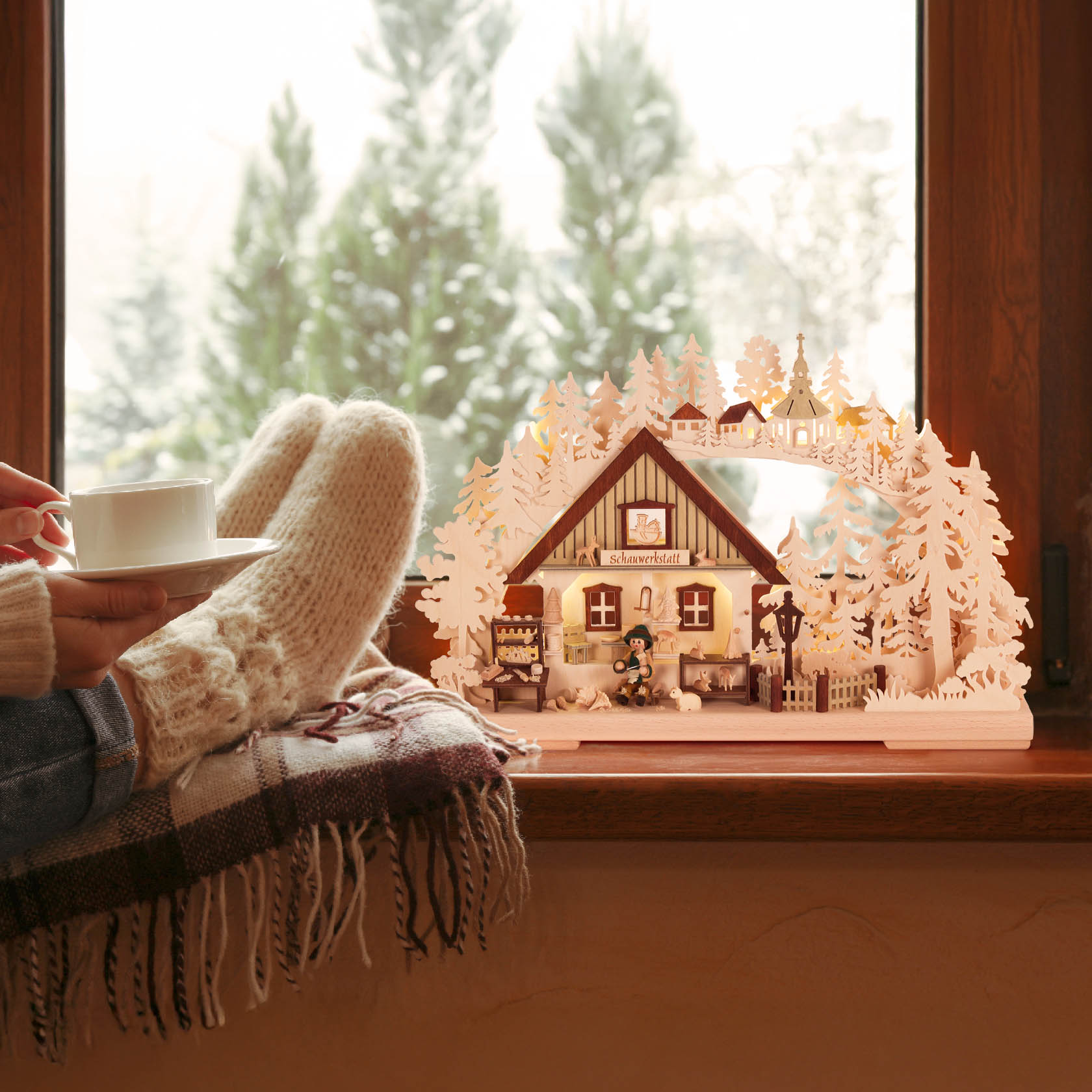 A small, electrically lit Schwibbogen depicting a workshop scene with a woodcarver. It's indoors, with a person in cozy socks holding a cup.