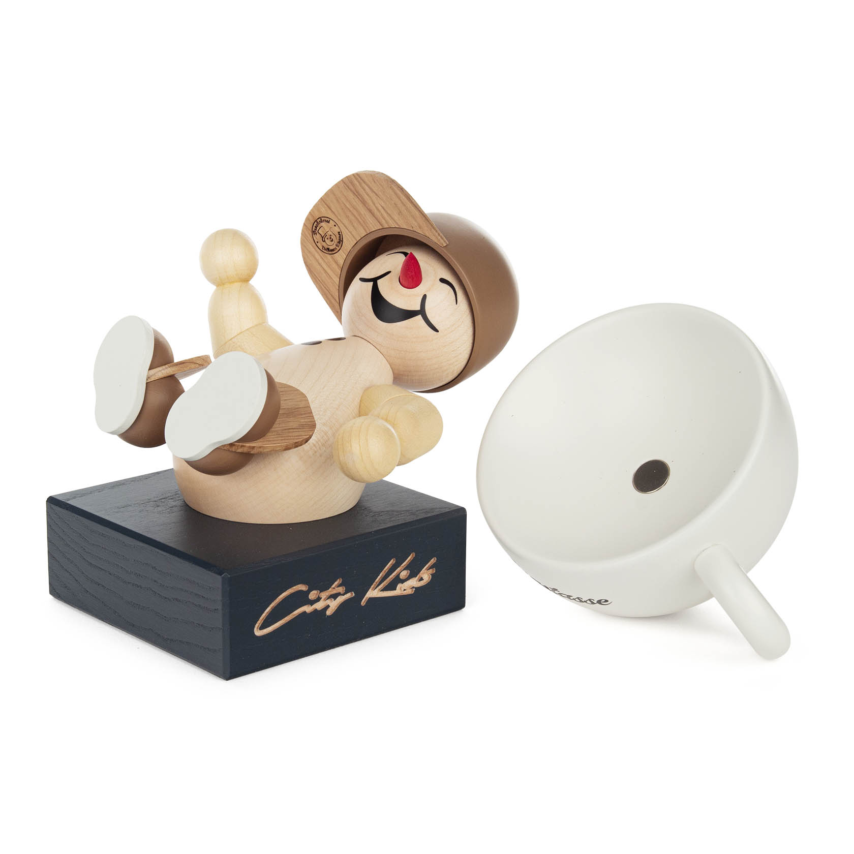 Wooden figure of a smiling child lying on its back, wearing a sideways cap, on a black base signed 'City Kids'. Beside it, a tilted white cup.