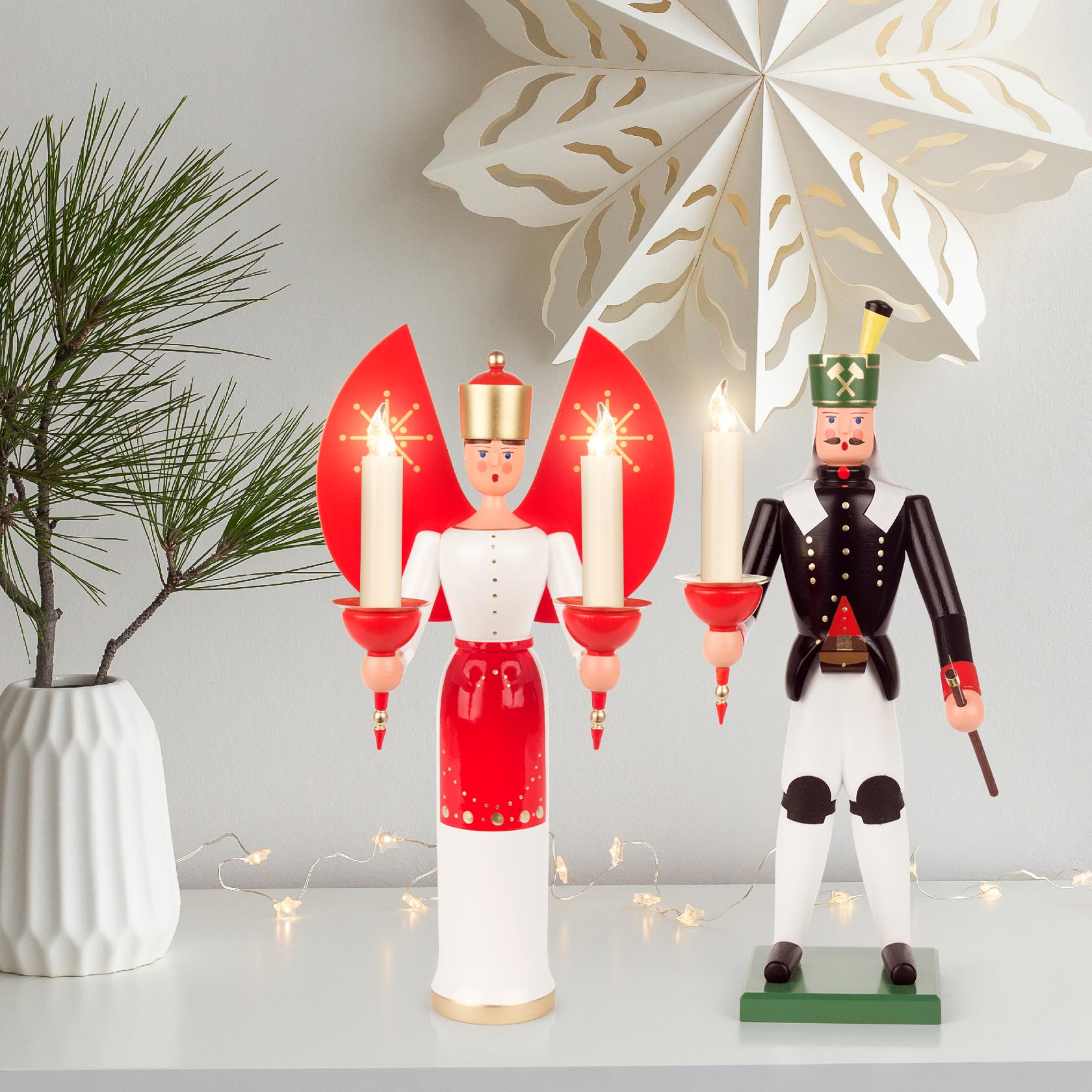 A wooden Christmas figure of a miner holding a candle holder designed for a 14mm candle, standing next to a wooden angel holding two red candle holders with lit candles.
