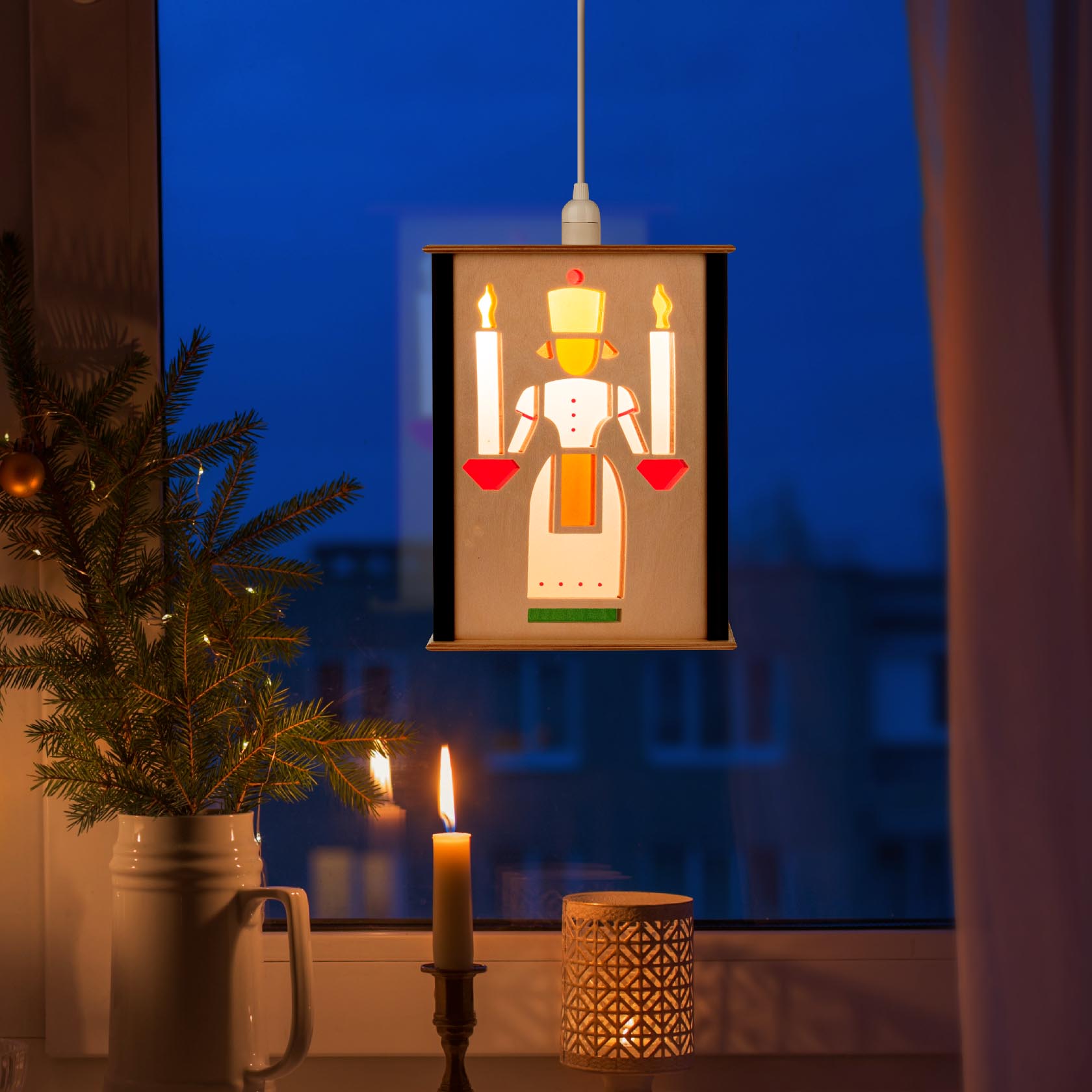 Fensterlaterne mit dem Motiv eines Lichterengels, der zwei Kerzen hält. Die Lampe hängt vor einem Fenster neben einem Tannenzweig mit einer Kerze im Vordergrund.
