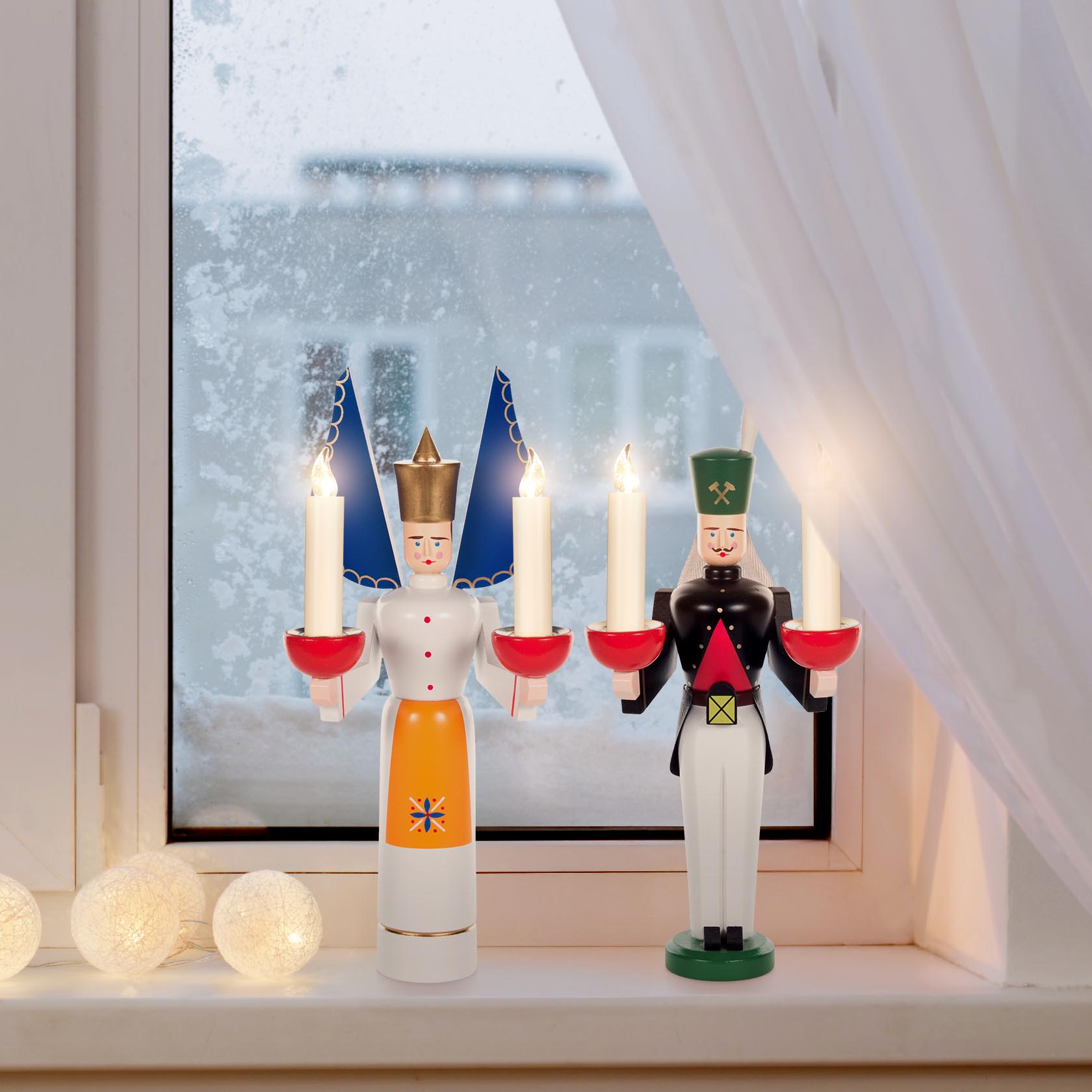 Traditionelle Holzfiguren Engel und Bergmann in Fensterdekoration. Engel mit blauer Flügel und Kerze, Bergmann mit grüner Mütze und Kerze.