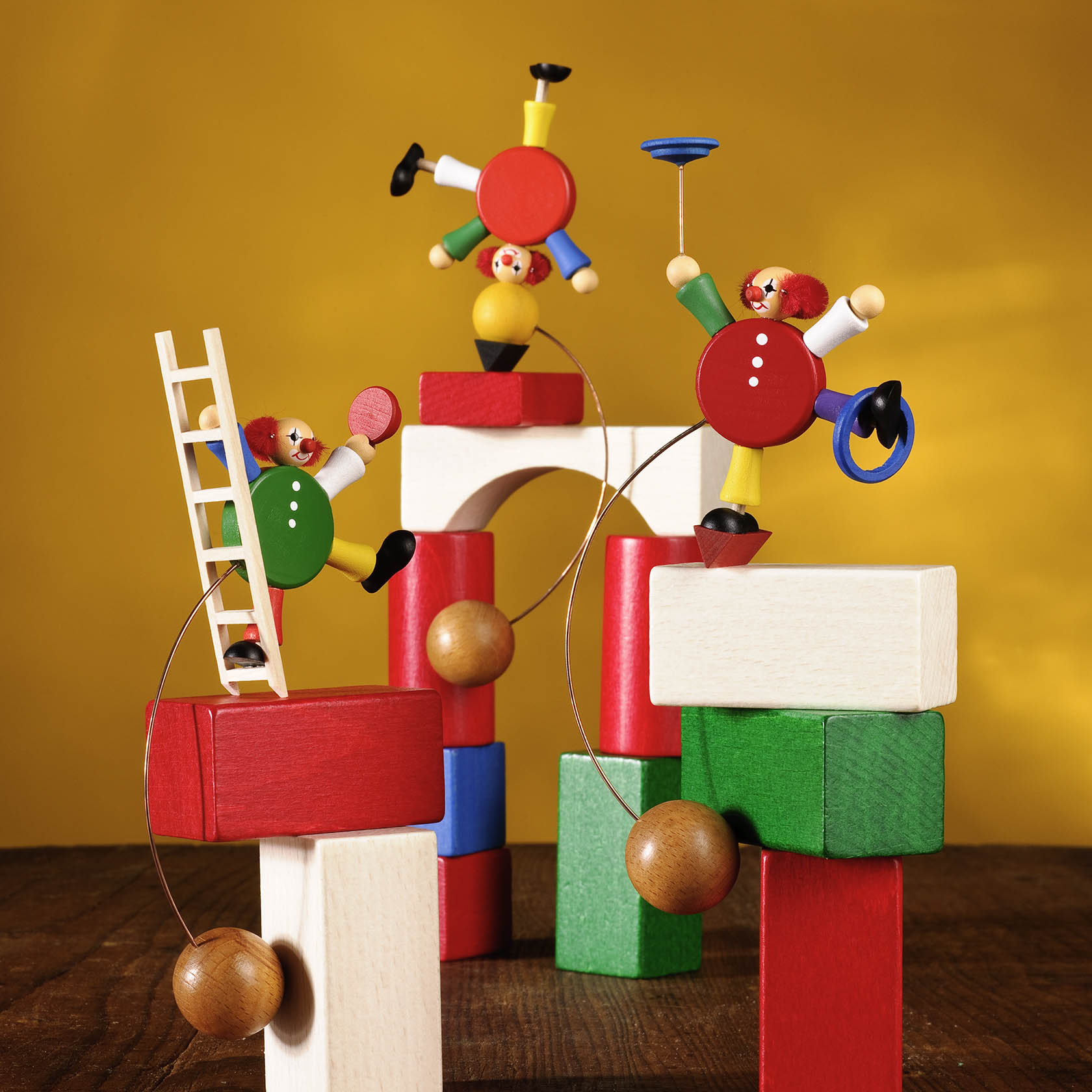 Drei bunte Holz-Clownfiguren balancieren akrobatisch auf Bauklötzen. Einer jongliert, einer balanciert mit Teller, der dritte klettert eine Leiter hoch.