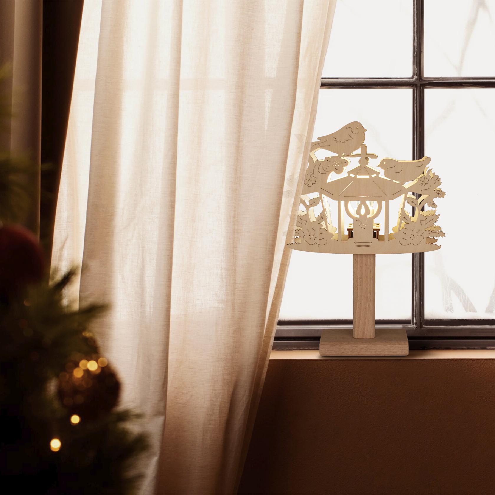 Ein Fensterbild zeigt eine Holzlaterne mit Vögeln und Blumen auf einem Sockel. Die Laterne ist elektrisch beleuchtet und im Fenster platziert.