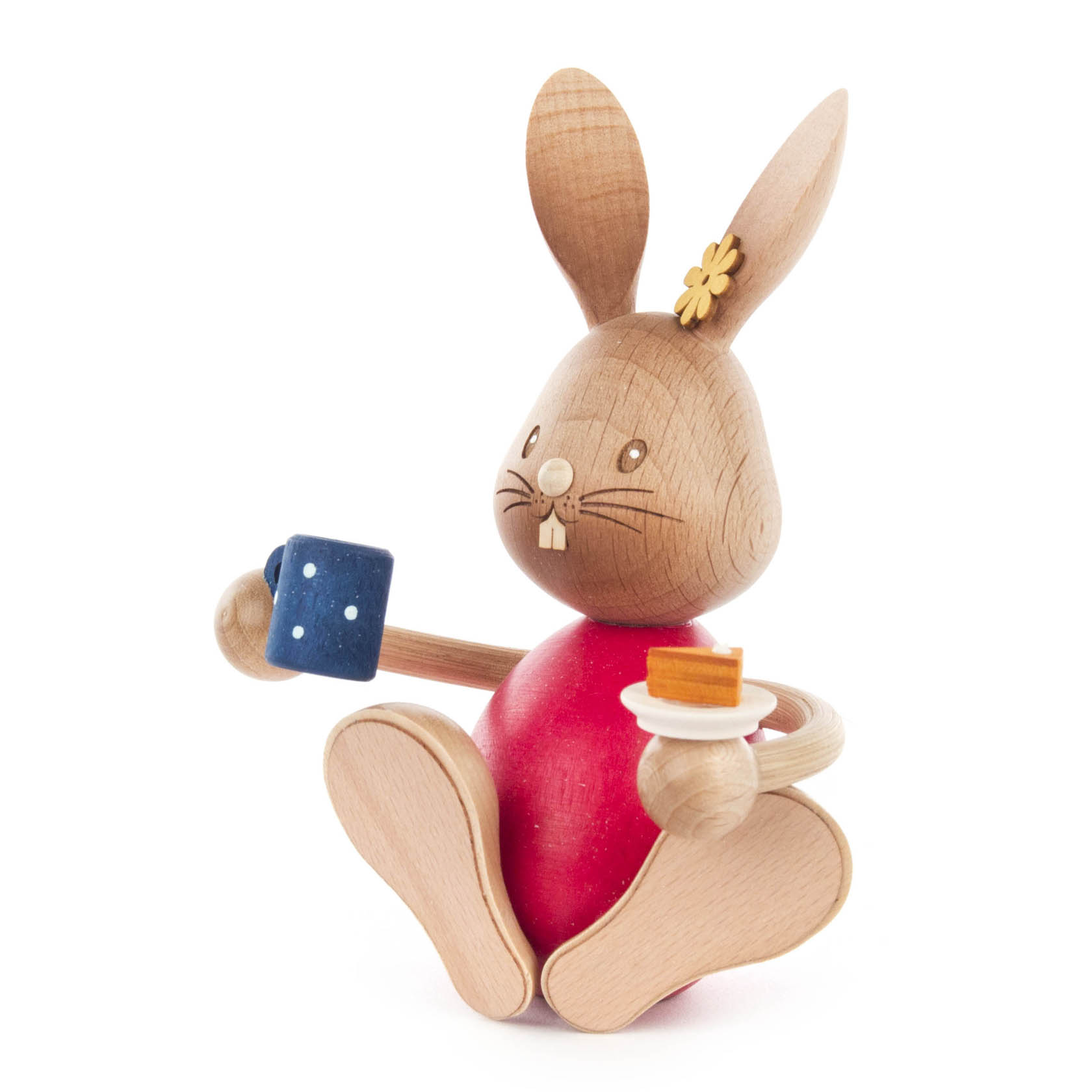 A wooden toy rabbit named Stupsi holding a blue coffee mug in one paw and a small plate with cake in the other, wearing a red shirt.