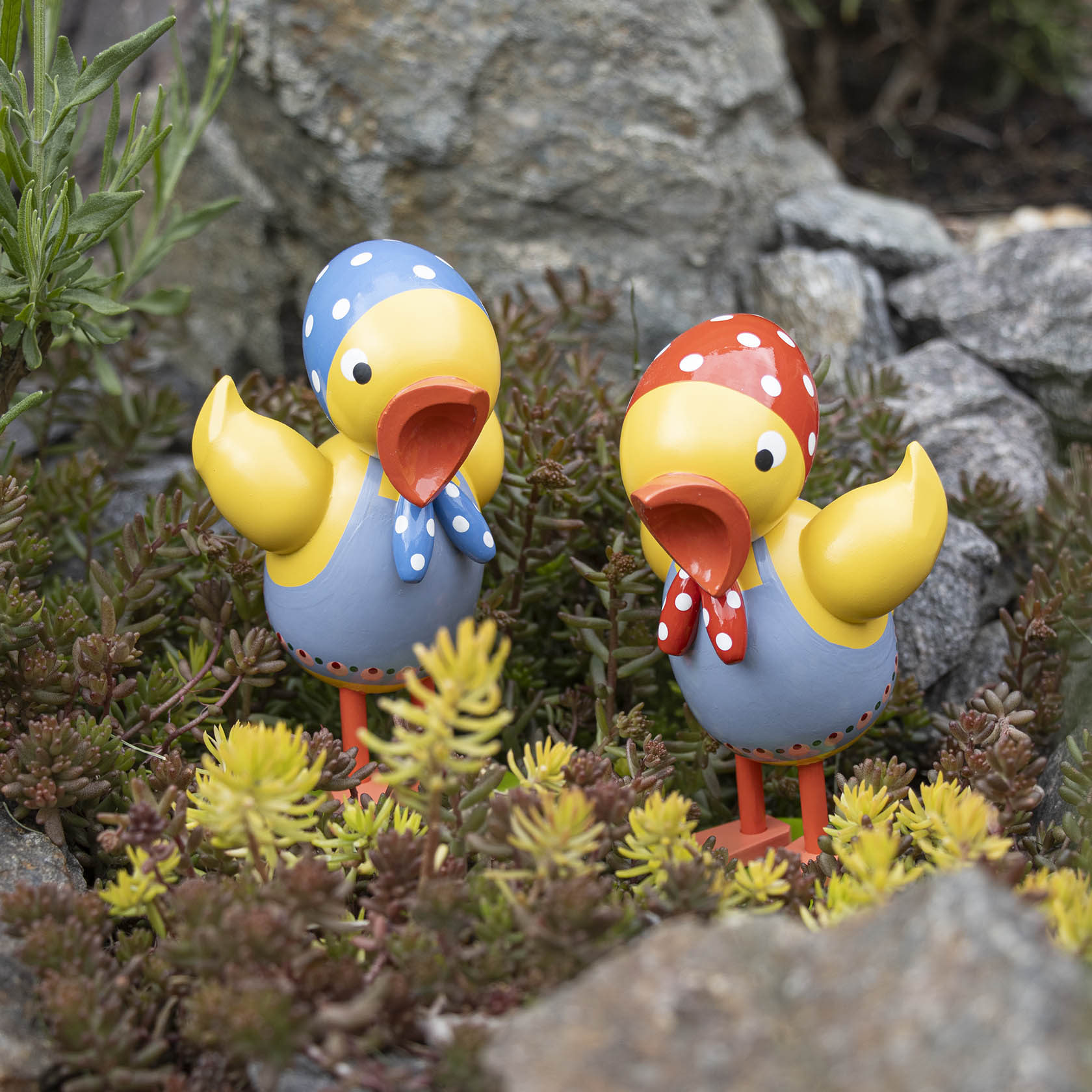 Zwei gelbe Kükenfiguren mit bunten Kopftüchern und Schürzen stehen in einer Gartenlandschaft.