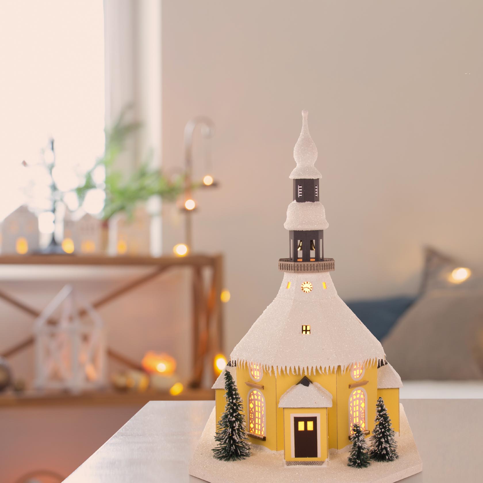 Ein detailreiches Modell der Seiffener Kirche mit elektrischer Beleuchtung, gelbem Gebäude, schneebedecktem Dach und kleinen Tannenbäumen vorn.