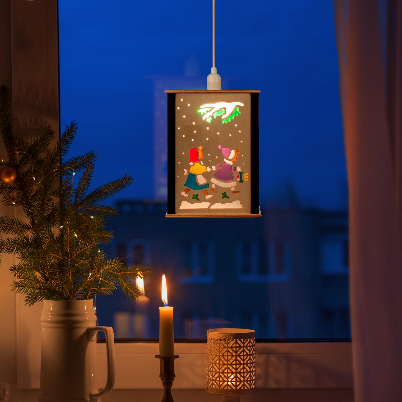 Eine kleine Fensterlaterne mit Kinder- und Wintermotiv, eingeschaltet und von innen beleuchtet, hängt vor einem Fenster.