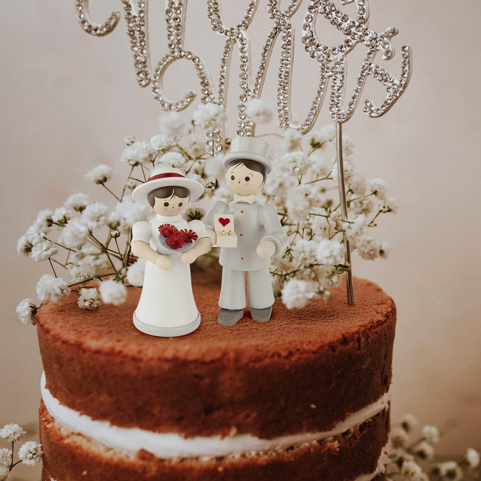 Colorful bridal figurines on a cake, featuring a bride in a white dress and hat holding a bouquet, and a groom in a light suit holding a card.