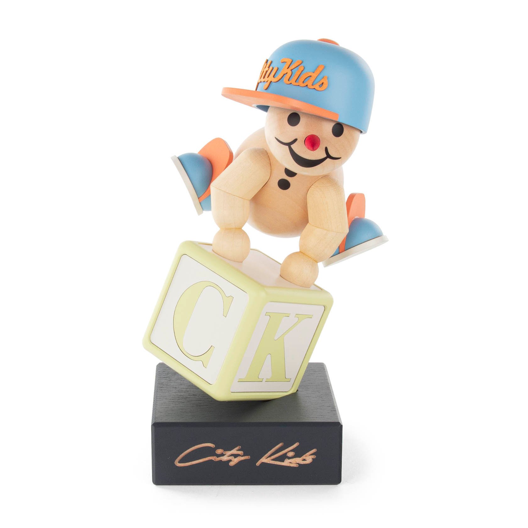 Holzfigur eines Kindes mit Baseballkappe, das auf einem Würfel mit den Buchstaben C und K balanciert. Die Base hat die Aufschrift 'City Kids'.
