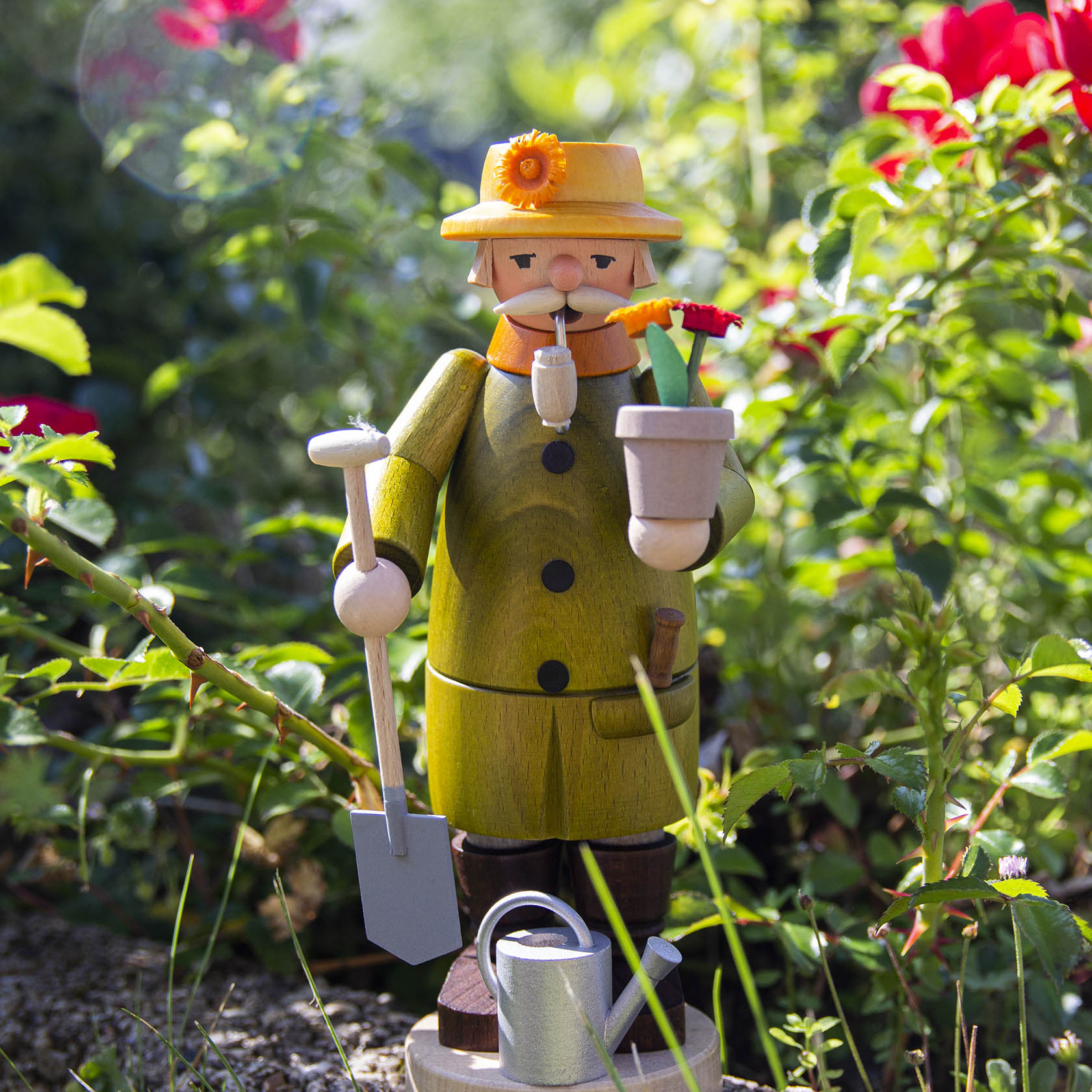 A wooden smoking figure of a gardener holding a flower pot with red flowers, wearing a yellow coat and hat, standing with a shovel and watering can nearby.