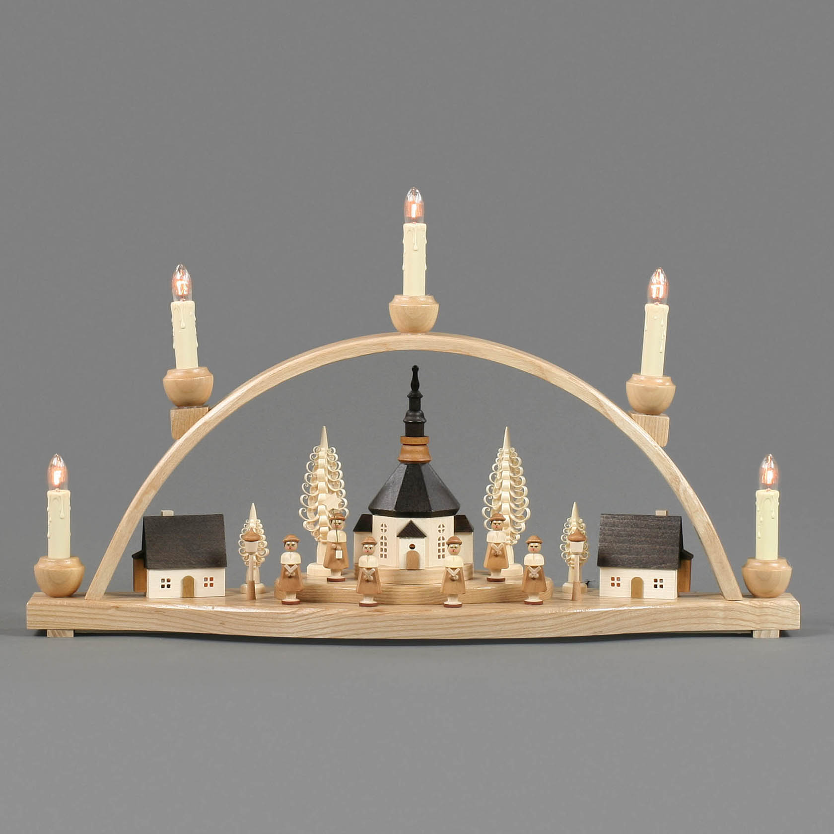 Wooden arch decoration with electric candles, depicting a Seiffen village scene with a church, trees, and choir figures, buildings internally lit.
