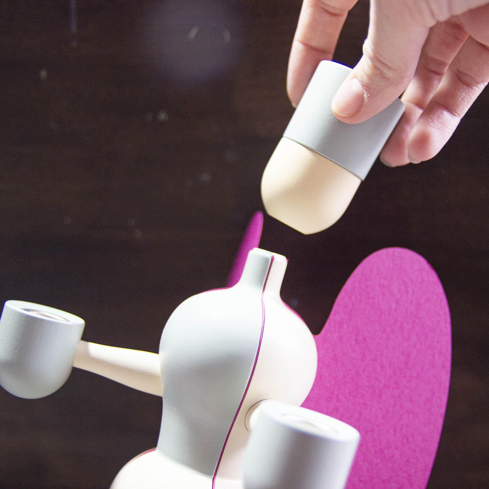 Holzengel mit zwei weißen Kerzenhaltern, eine Hand fügt zylinderförmigen Kopf hinzu. Rosa Flügel im Hintergrund.