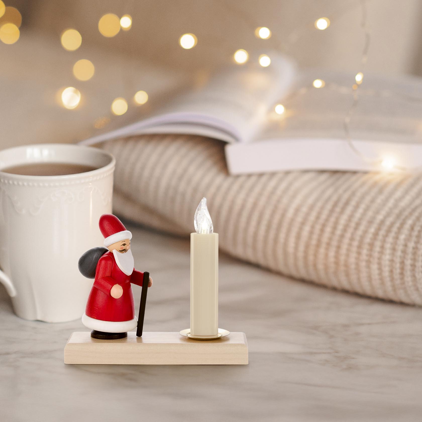Ein Holzkerzenhalter zeigt Ruprecht in rotem Mantel mit Stock und Sack neben einer Kerze. Hintergrund ist unscharf mit Lichterkette und Tasse.