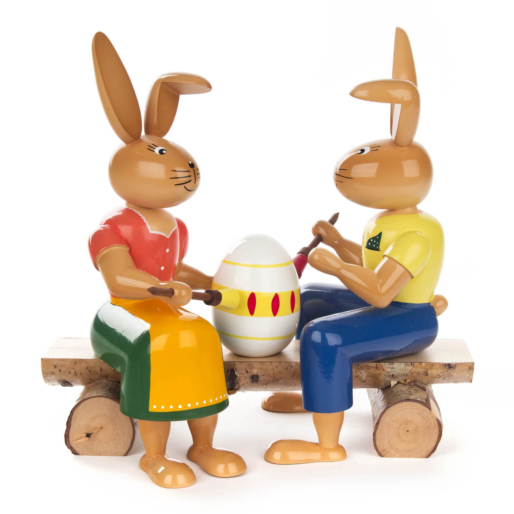 Two painted wooden rabbits sitting on a bench, each holding a paintbrush, with an Easter egg between them, dressed in traditional clothing.