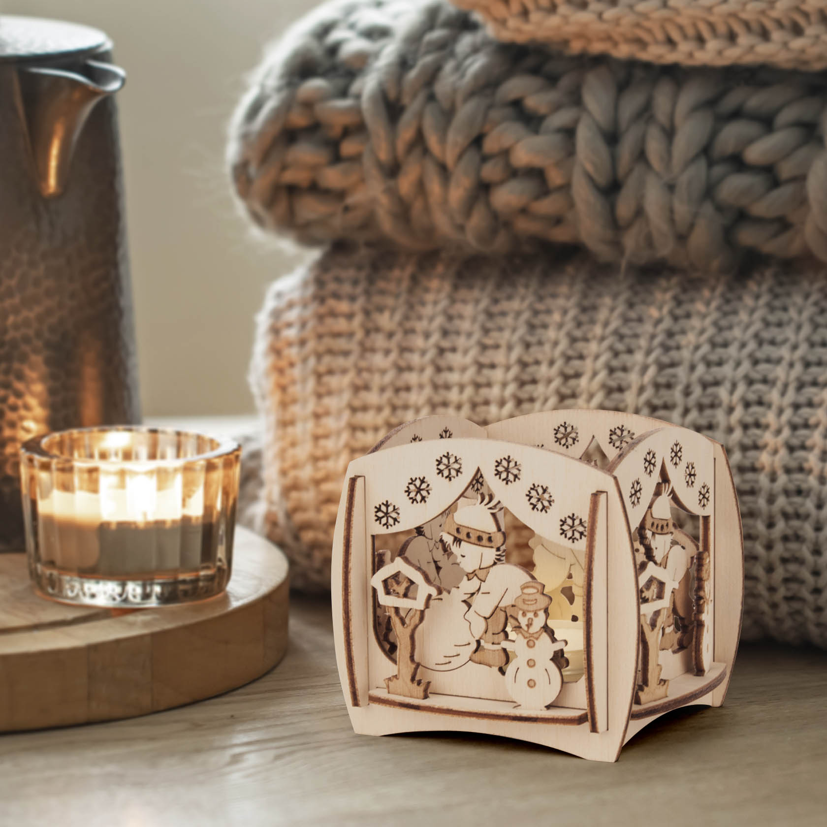 A wooden tealight holder with a winter scene featuring snowmen and snowflakes, surrounded by cozy blankets and a lit candle.