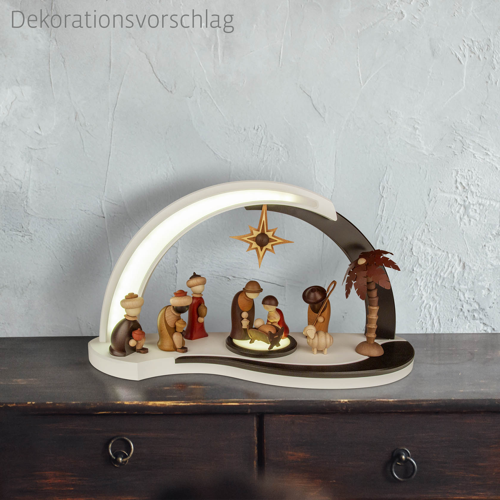 Neunteilige Krippefiguren-Szene mit Maria, Josef, dem Jesuskind unter einem Stern und weiteren Figuren um eine Palme, auf einem dekorativen Bogen.