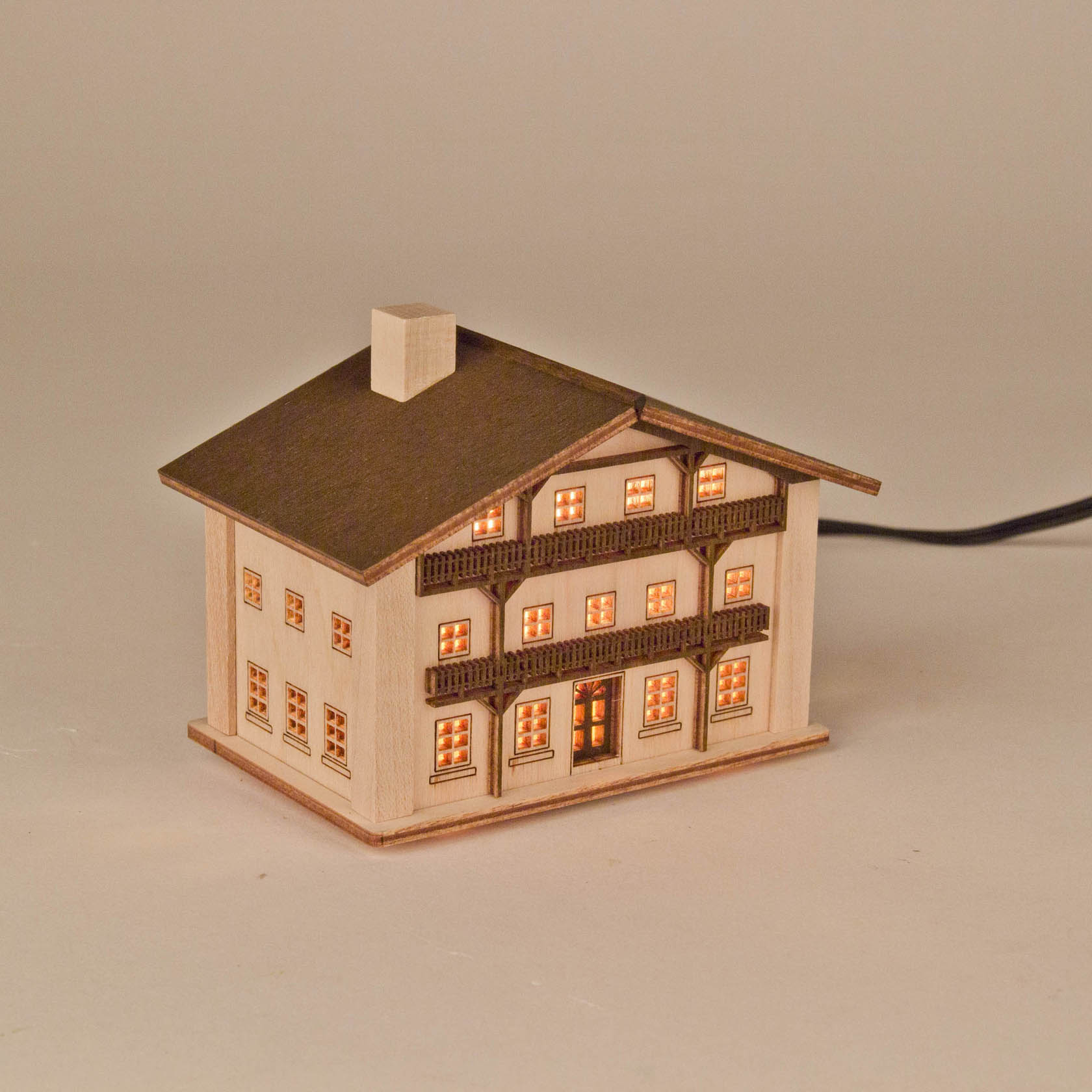 A wooden model of a mountain house illuminated from within, showcasing multiple glowing windows and a detailed balcony.
