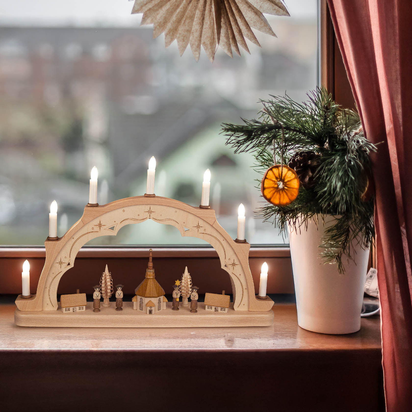 Ein Holz-Schwibbogen mit elektrischer Beleuchtung zeigt eine Kurrendeszene mit Figuren und Kirche auf einer Fensterbank.