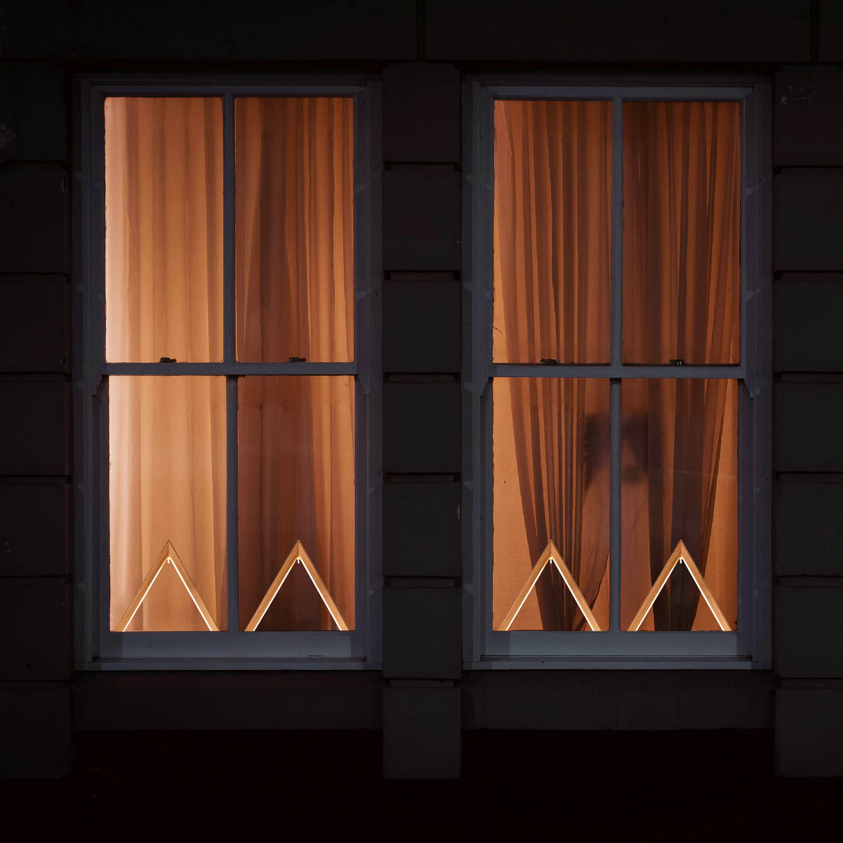 Zwei Fenster mit geschlossenen Vorhängen, dahinter beleuchtete dekorative Dreiecke mit LED-Band.