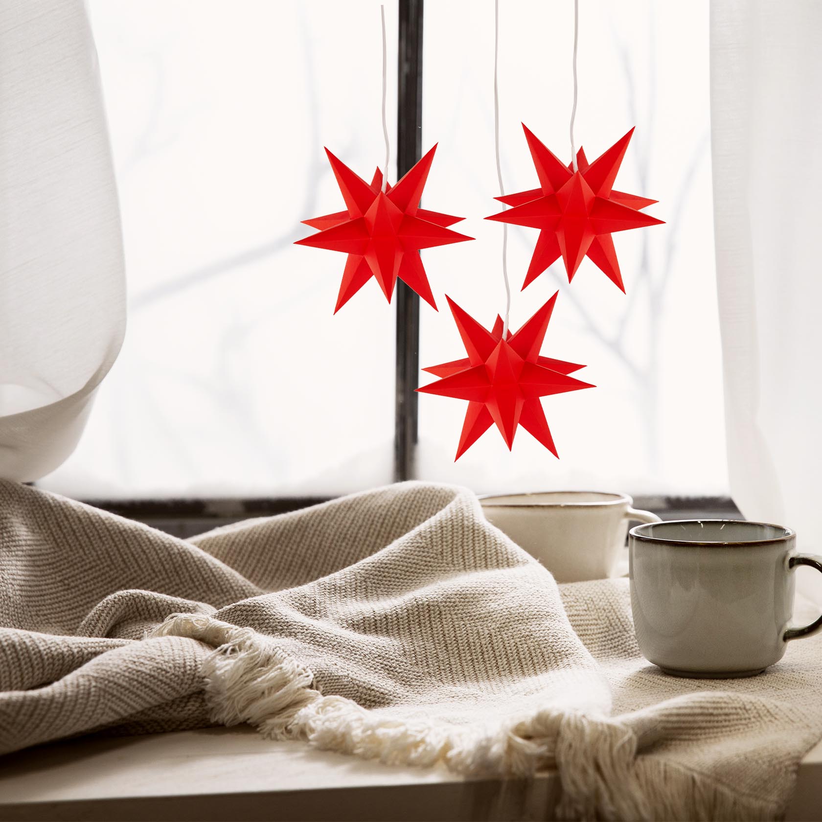 Three small red star-shaped paper lamps with electric lighting hanging by a window beside two ceramic mugs on a soft, textured cloth.