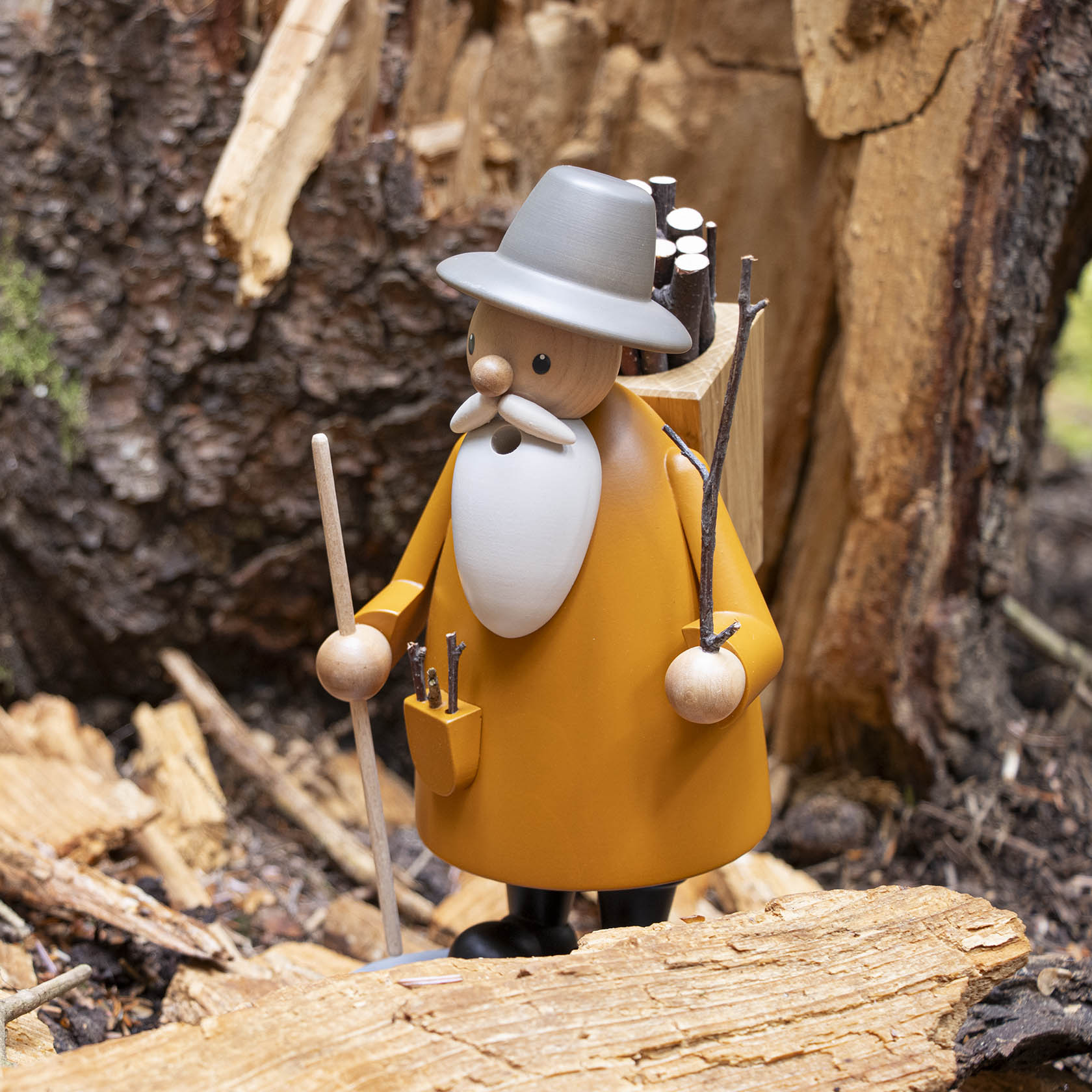 Holzfigur eines Mannes mit grauem Hut und großem weißen Bart, der Stöcke trägt, in einem Waldumfeld.