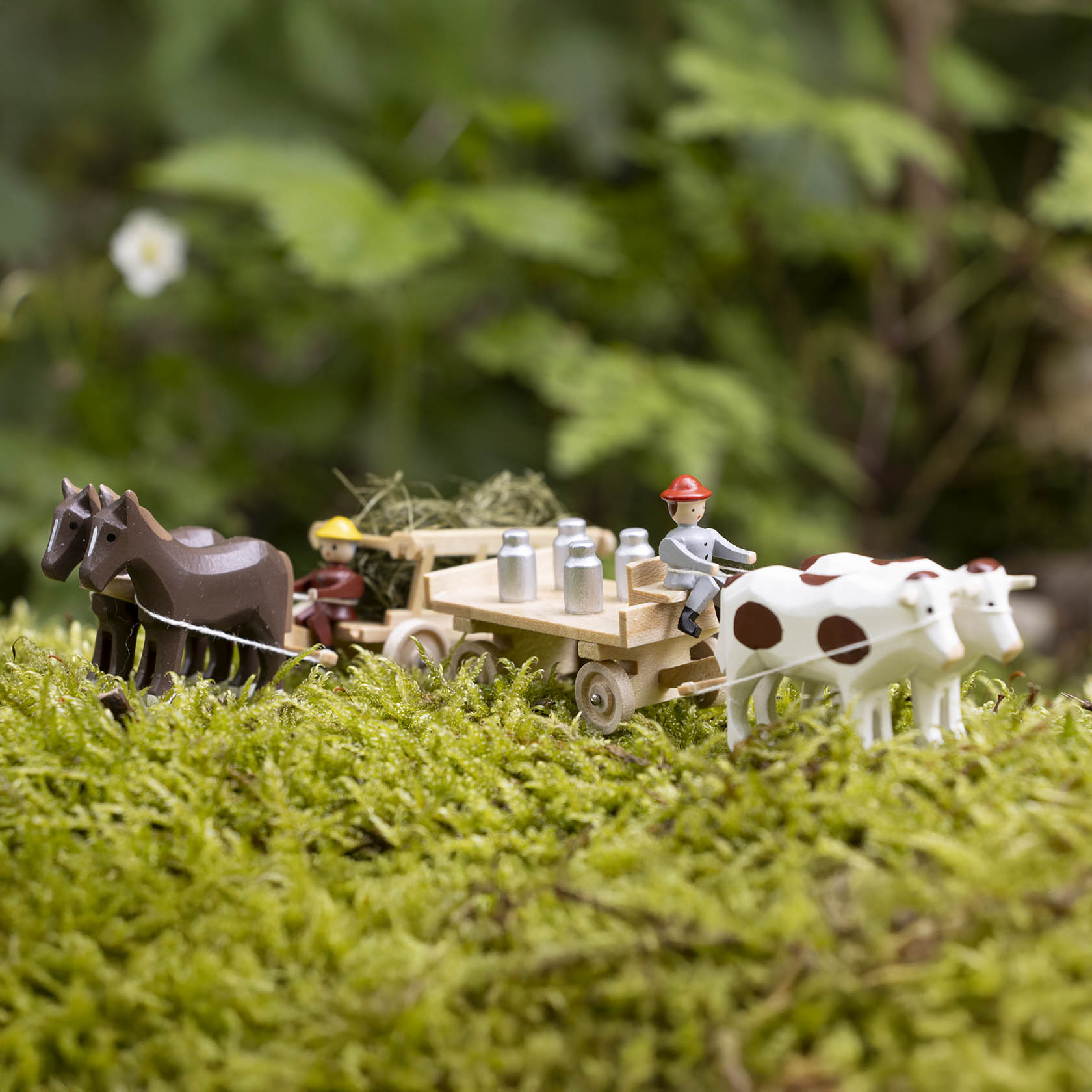 A model scene with horses pulling a wooden wagon carrying hay and milk cans. A figure in a hat leads the wagon, followed by two cows.