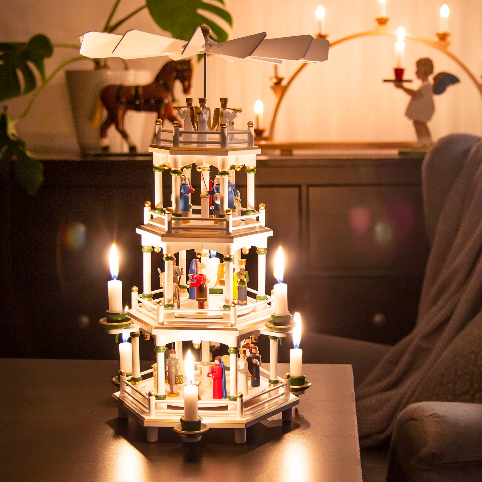 A three-tiered white Christmas pyramid depicting the Nativity scene, adorned with lit candles and figures, set on a cozy indoor table.