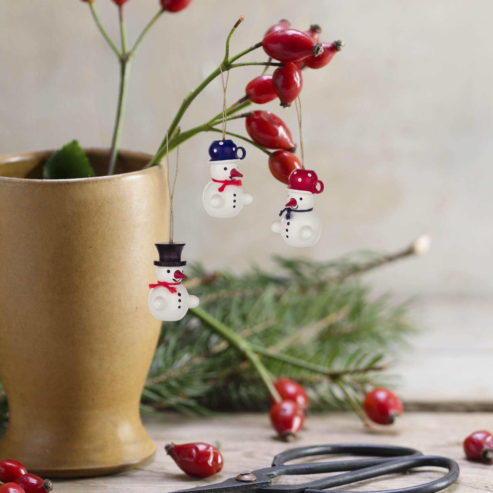 Three small snowman ornaments hang from a twig in a vase with red berries and green pine needles. Scissors are in the foreground.