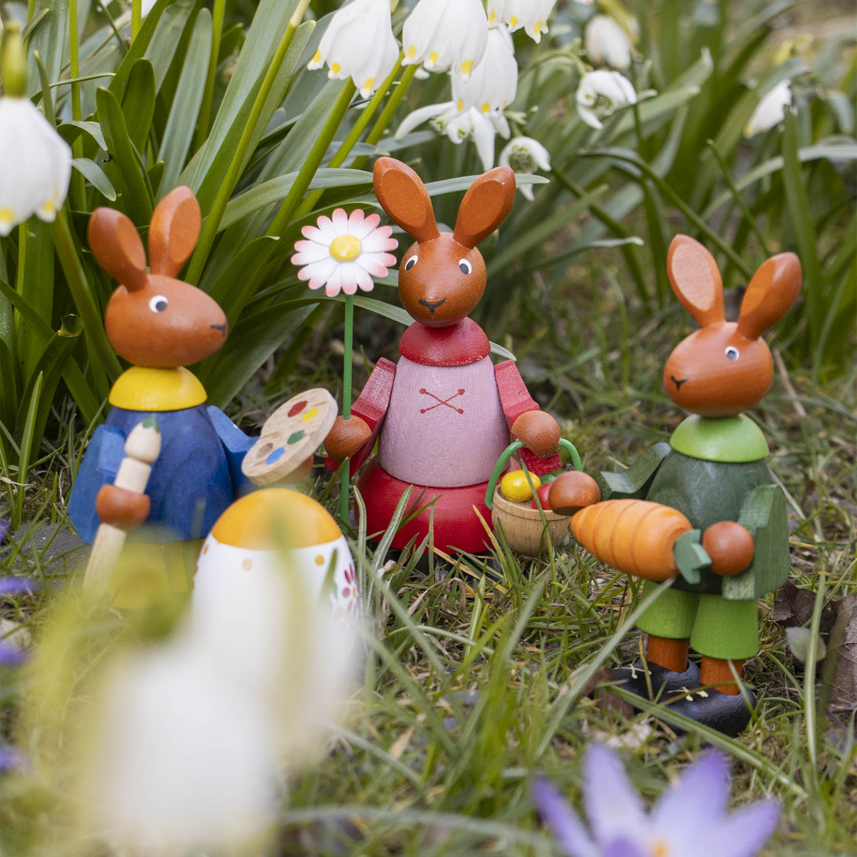 A group of four wooden Easter bunnies in colorful outfits, one holding a large carrot, standing on grass with spring flowers around.