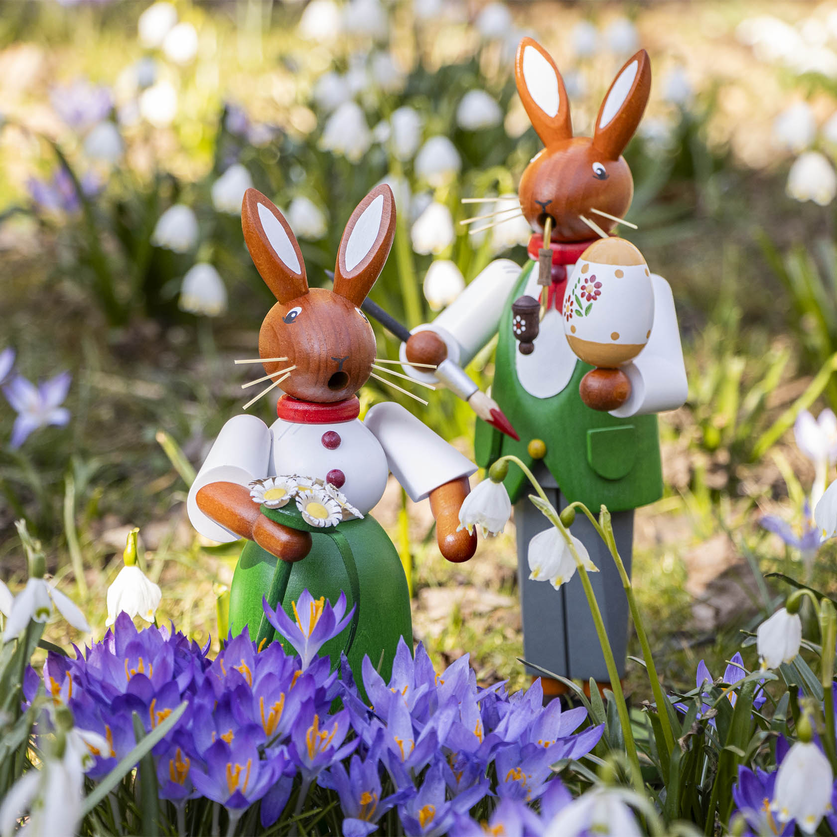 Zwei hölzerne Hasenfiguren, eine weiblich mit Blumenstrauß, die andere männlich mit Pfeife und Osterei, in einem Blumenfeld.