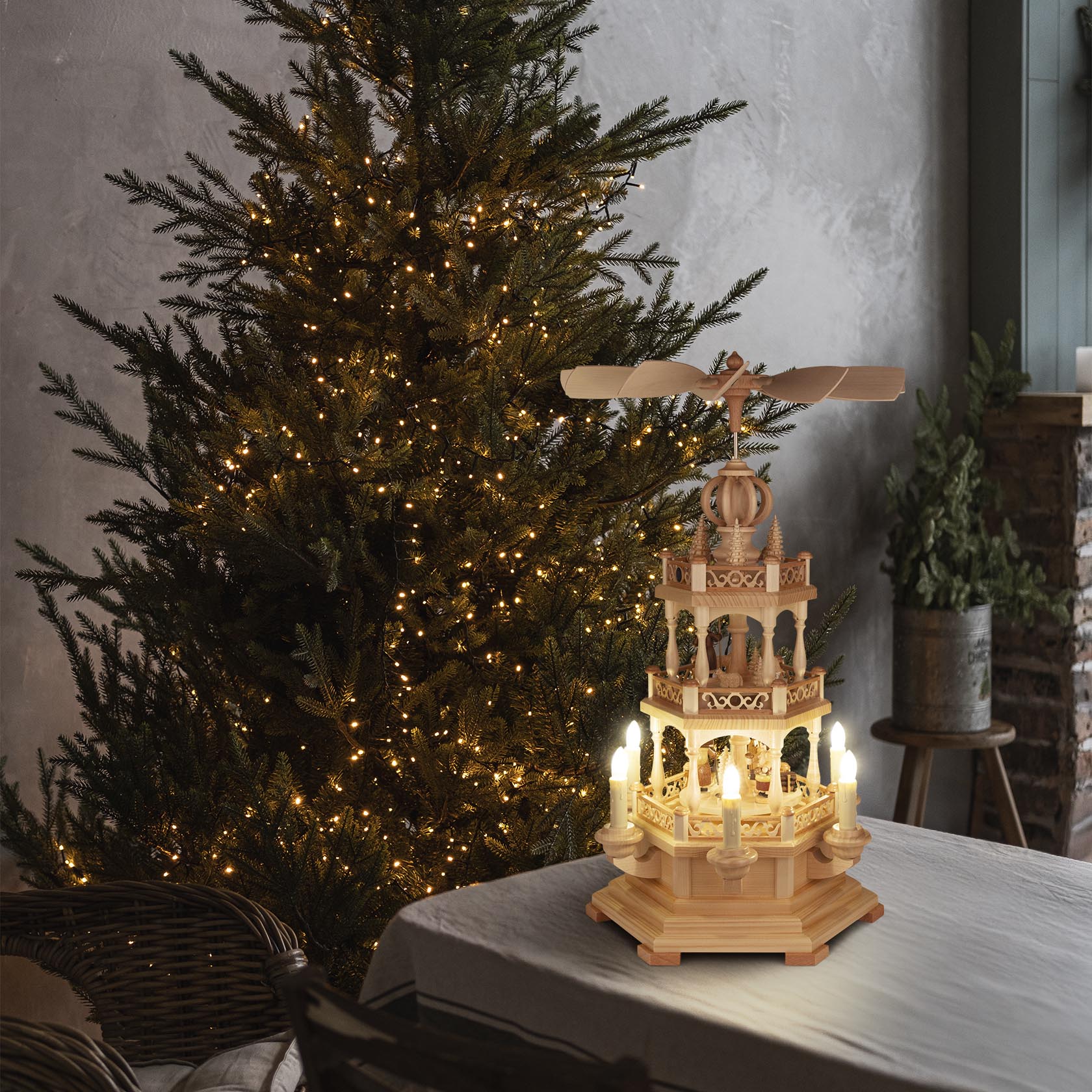 Zweistufige, hölzerne Weihnachtspyramide auf einem Tisch, umgeben von einem beleuchteten Weihnachtsbaum. Die Pyramide hat bewegliche Flügel oben und ist elektrisch beleuchtet.