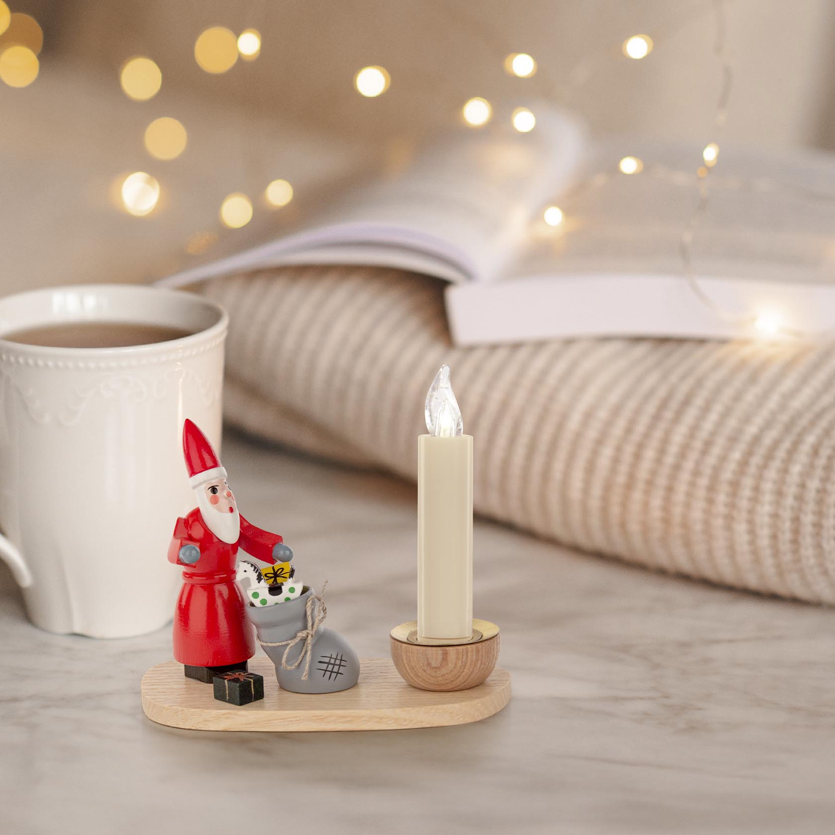 Ein Kerzenhalter mit einem Weihnachtsmann aus Holz, der ein Geschenk und einen Sack hält, mit einer LED-Kerze, auf einem Holztisch, daneben eine Tasse.