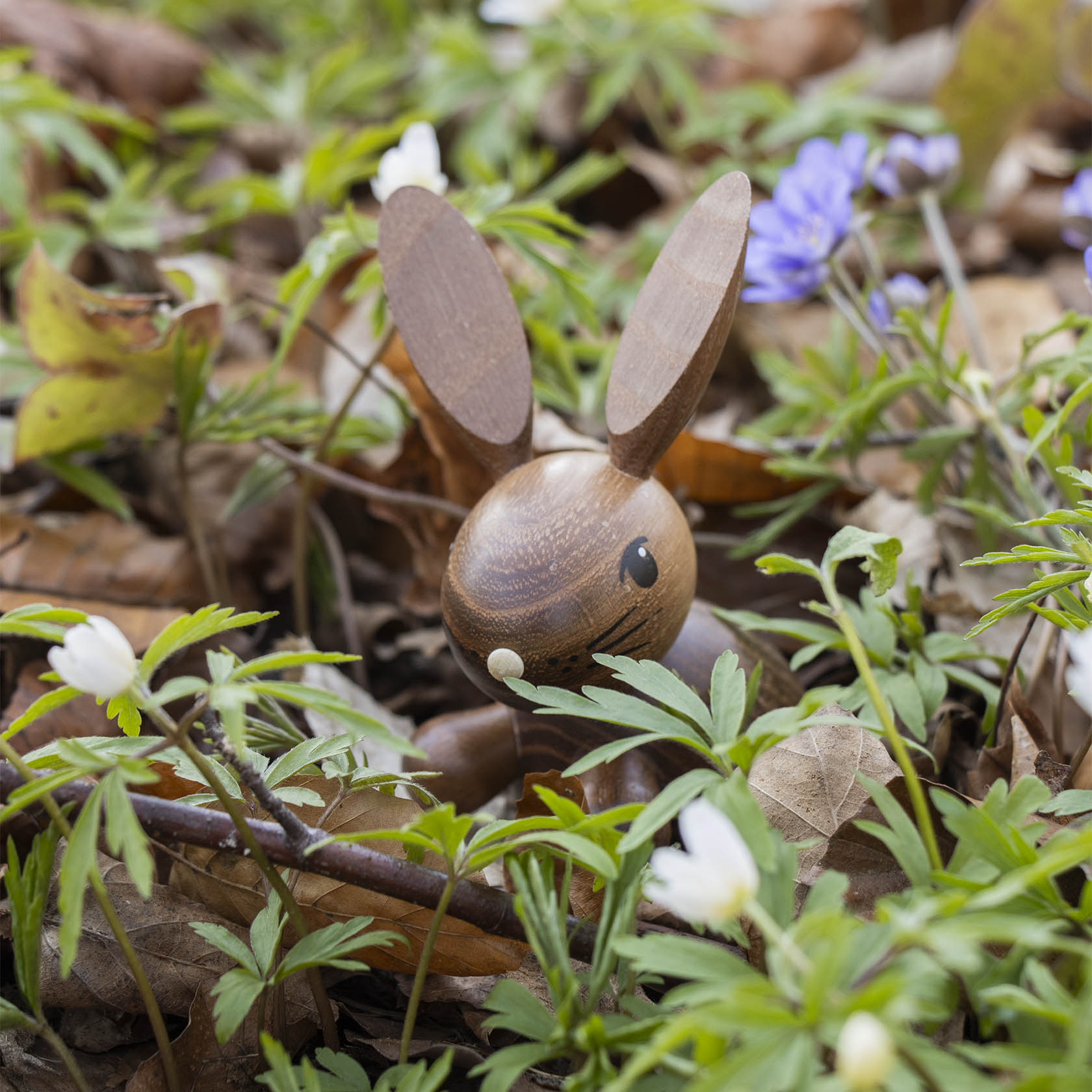 Ein dunkelbrauner Holz-Hase namens 'Henry' liegt im Laub und wird von kleinen weißen und blauen Blumen umgeben.