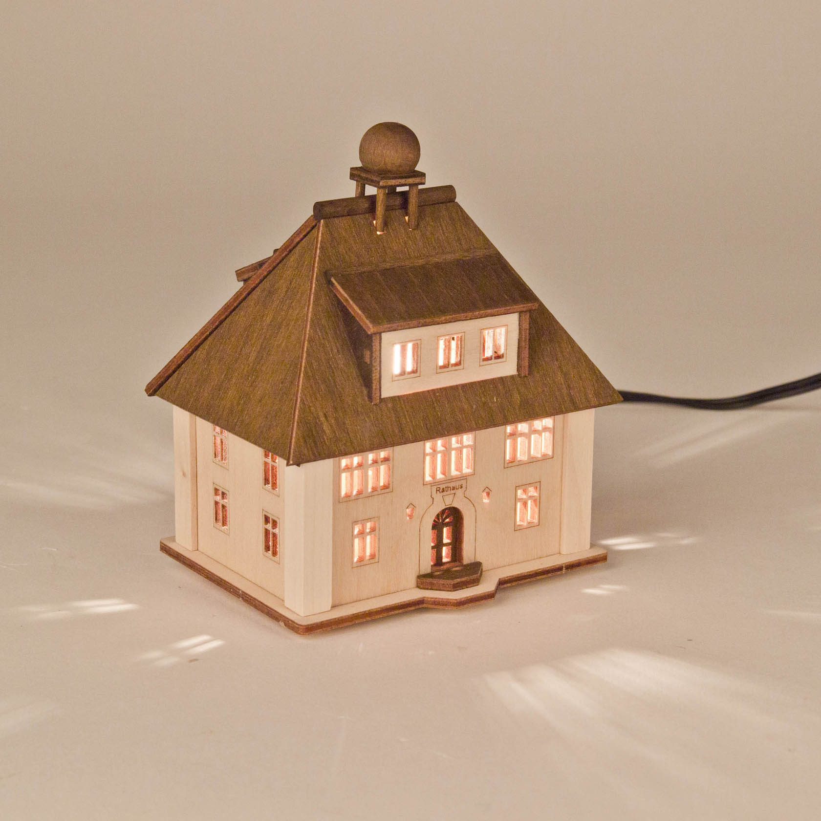 Ein kleines, dekoratives Holzhaus in Form eines Rathauses, mit erleuchteten Fenstern. Das Haus hat ein großes Dach mit einer Kugel oben drauf.