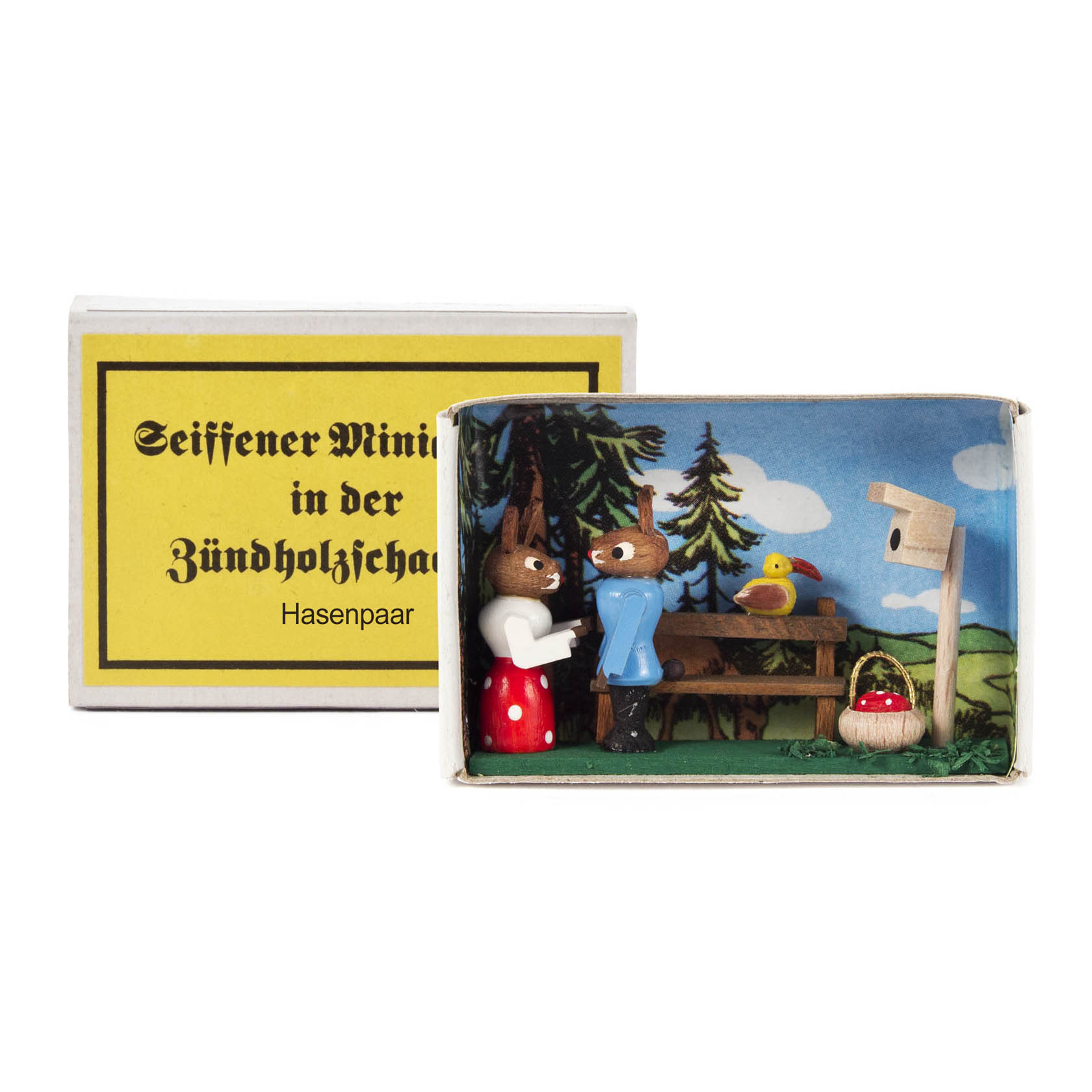 Two handcrafted wooden bunnies inside a matchbox, one wearing a red skirt and the other a blue jacket, beside a table with a basket and a birdhouse.