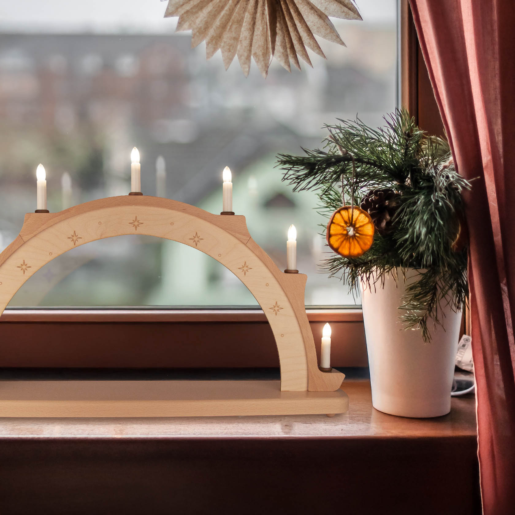 A wooden arch with electric candles on a windowsill, unadorned, next to a pot with pine branches and an orange slice.