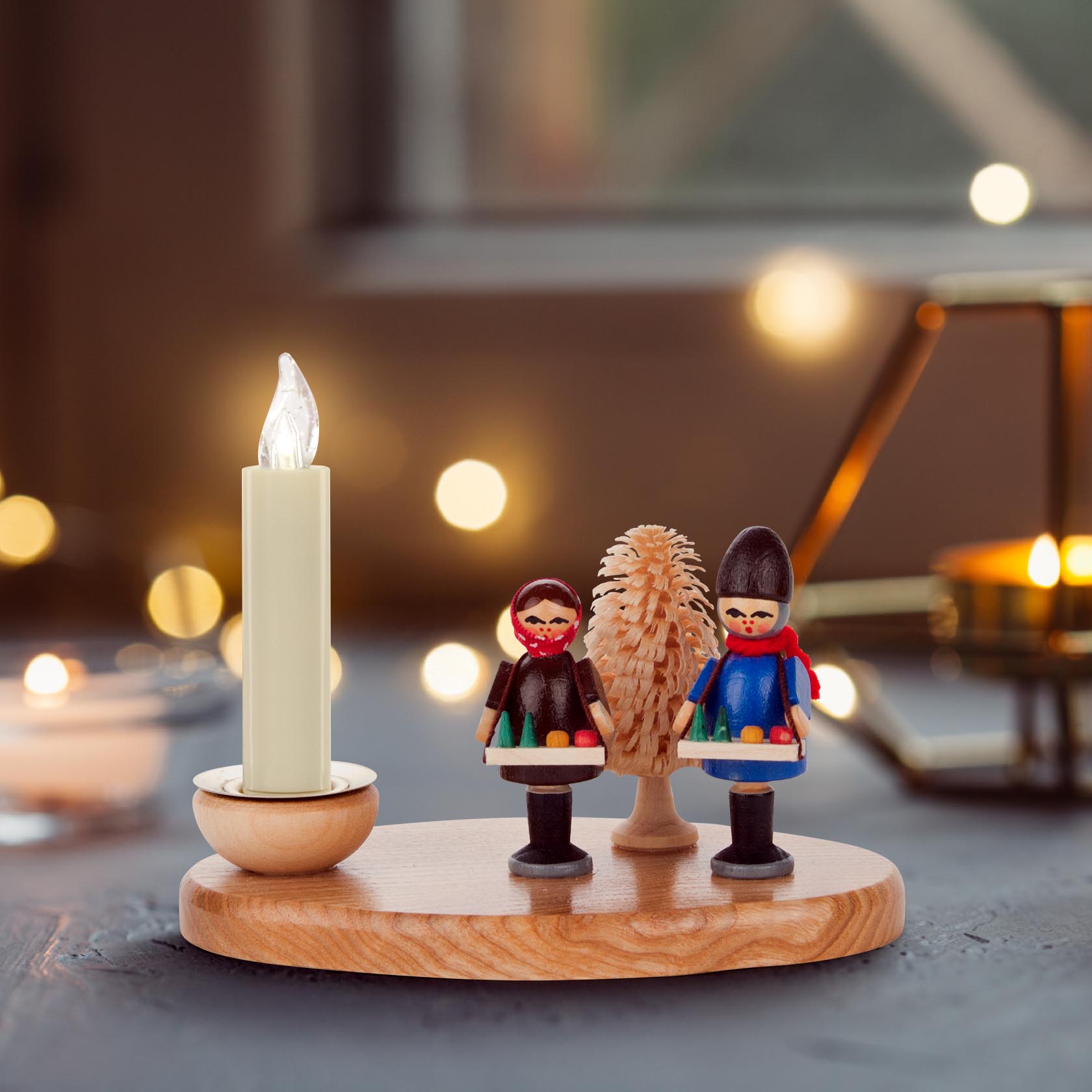 Holzkerzenhalter mit zwei bemalten Figuren, die traditionell gekleidet sind, eine brennende Kerze in der linken Halterung, mittig ein Holzbaum.