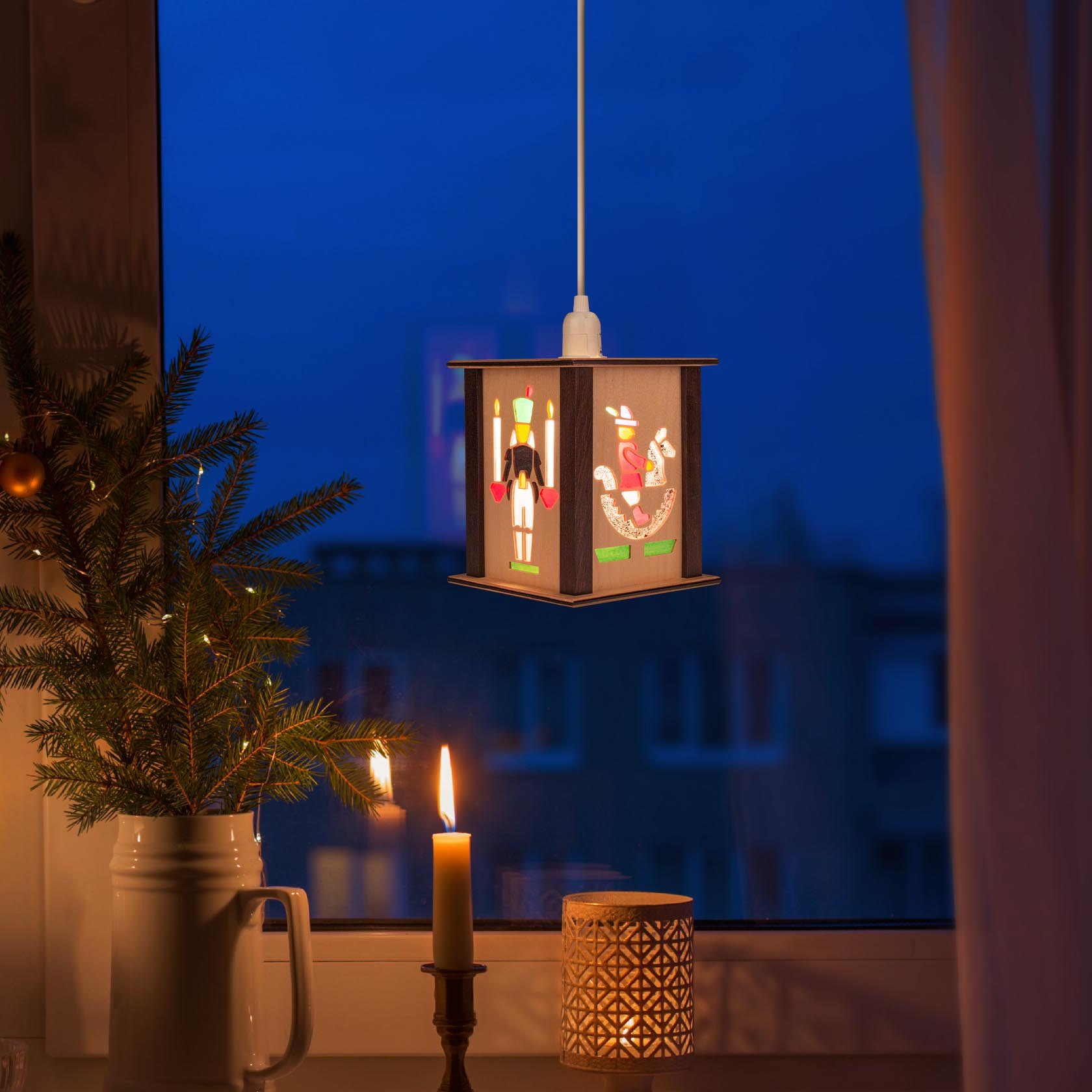 A small lantern with images of a rider and a horse, hanging indoors by a window next to a lit candle and a fir branch.