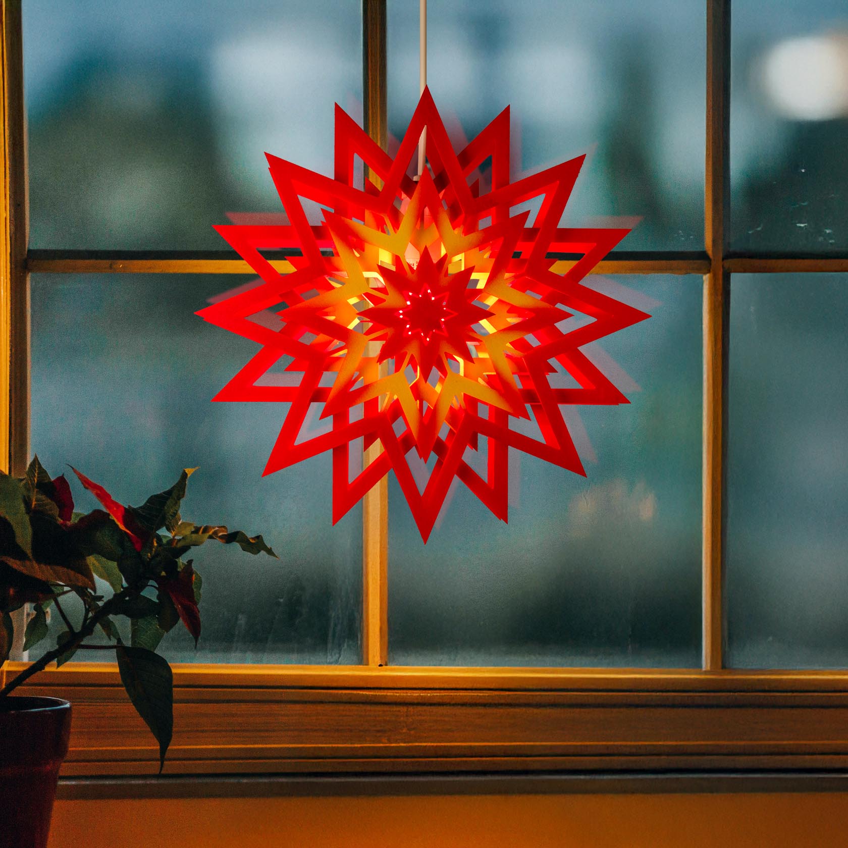 Illuminated red and yellow paper star hanging in a window, glowing brightly as the evening sets in.