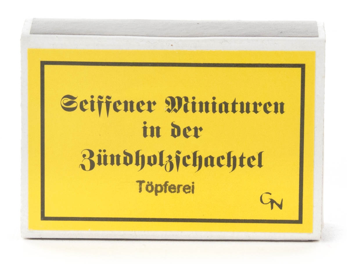 Zündholzschachtel mit gelbem Etikett, Aufschrift: 'Seiffener Miniaturen in der Zündholzschachtel Töpferei' und Initialen 'GN'.