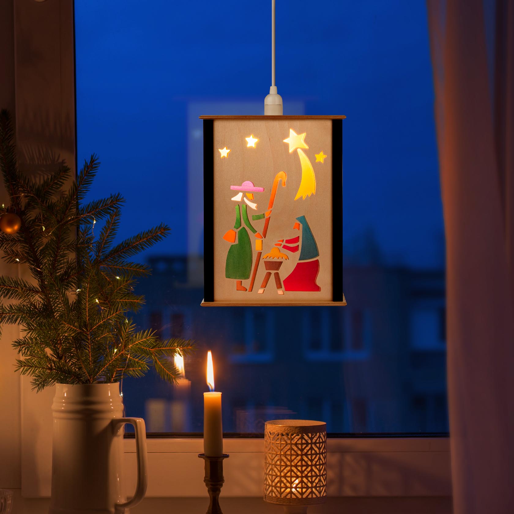 Beleuchtete Laterne im Fenster zeigt die Geburt Christi mit Maria, Josef und dem Jesuskind im Stall, begleitet von einem Stern und leuchtenden Kerzen.