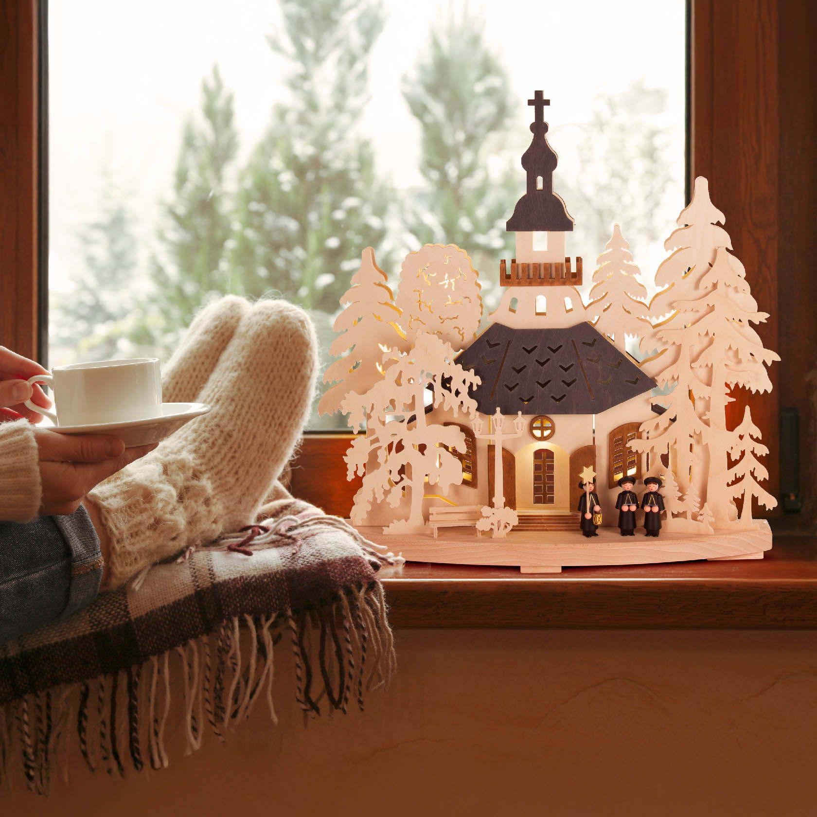A cozy scene with a person in wool socks holding a cup next to a lit wooden arch depicting a church with figures in winter attire, trees, and glowing windows.