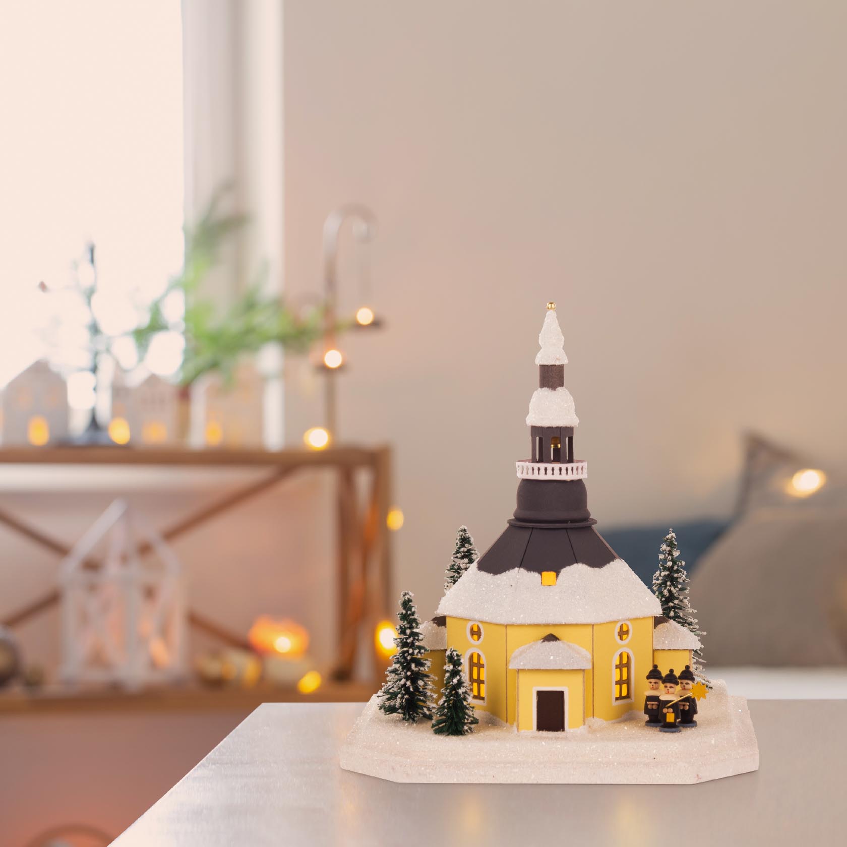 A small decorative Seiffen church model with electric lighting, featuring a snow-covered roof, surrounded by miniature evergreen trees and subtle interior lighting.