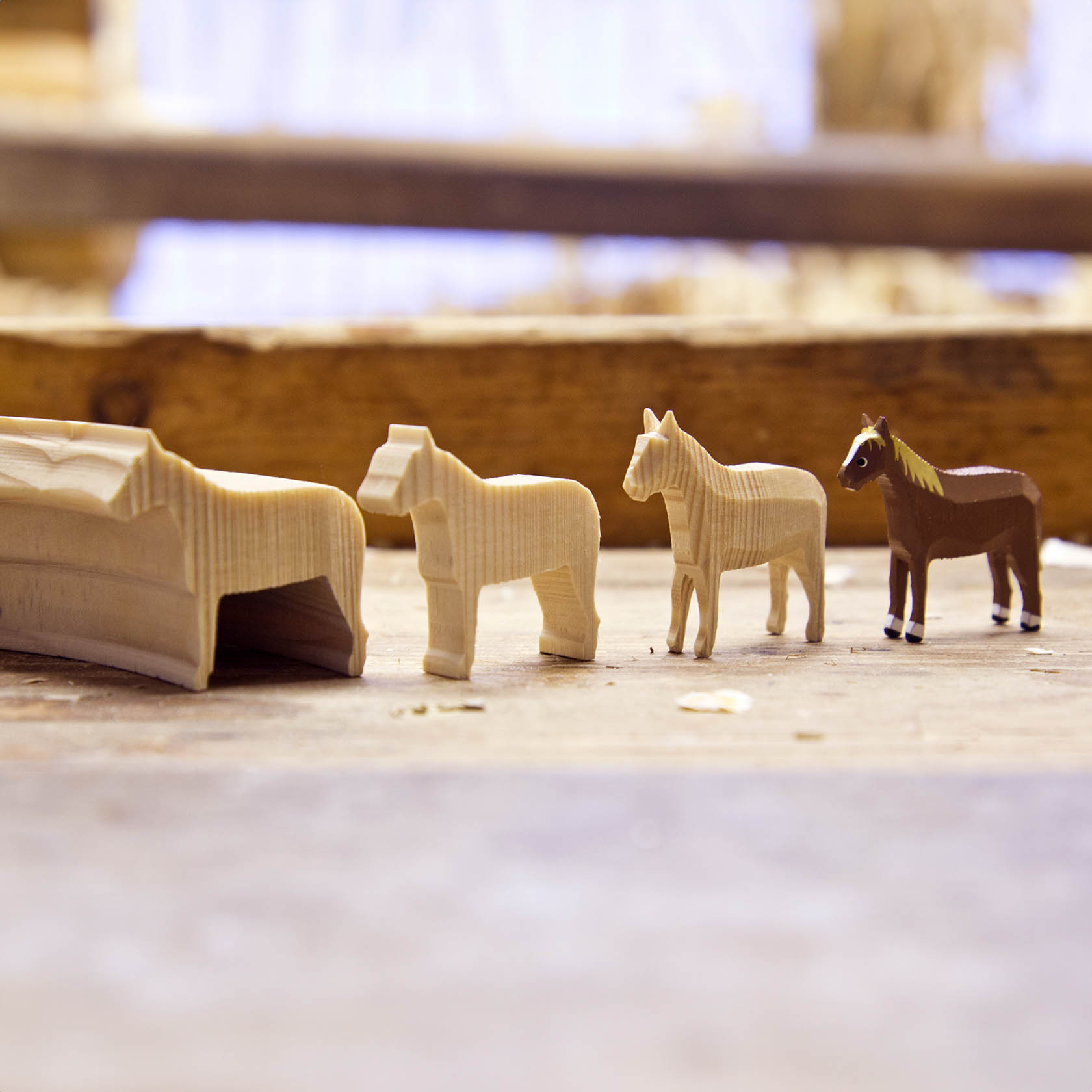 Four wooden animal figures, including different types of horses, lined up on a wooden surface next to a small wooden structure.