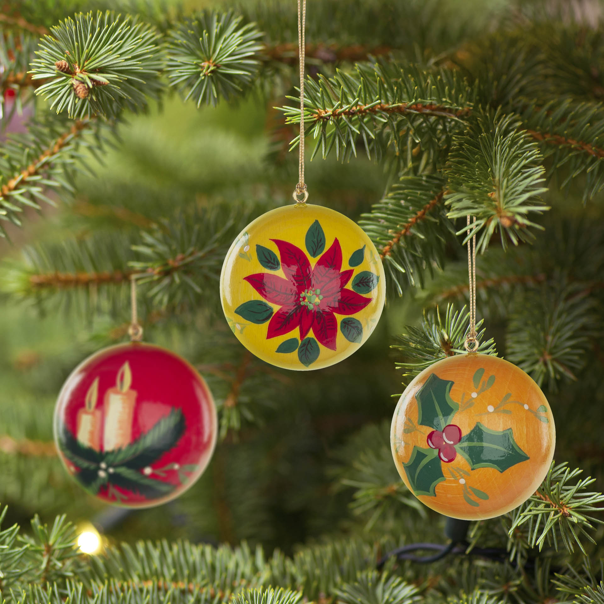 Three Christmas ornaments hang on a pine tree branch, featuring designs of candles, a poinsettia flower, and holly leaves in red, yellow, and orange colors.