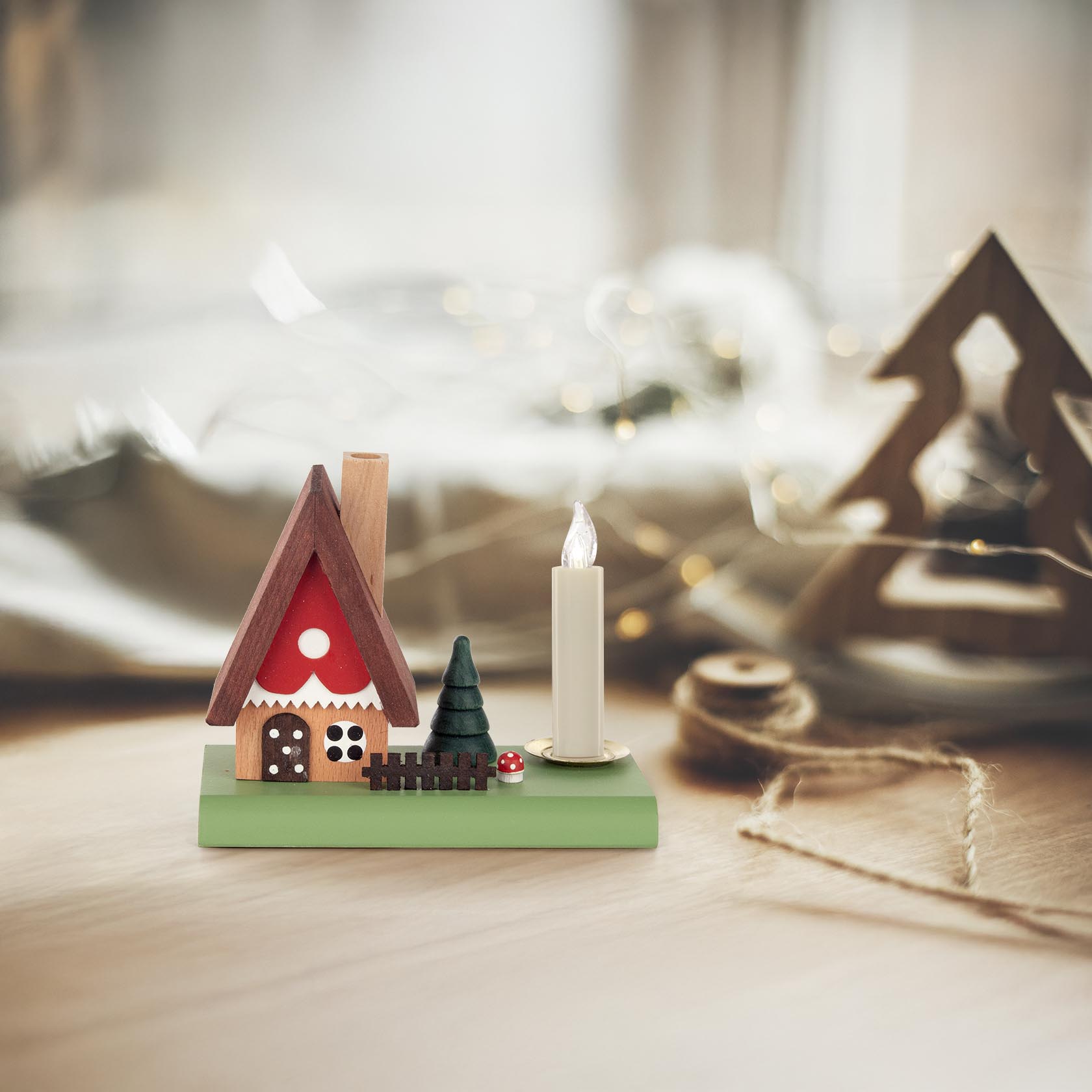 Kleine Holz-Nachbildung eines Märchenwaldhauses mit rotem Dach und zwei pilzförmigen Verzierungen, daneben brennende Kerze und ein kleiner Tannenbaum.